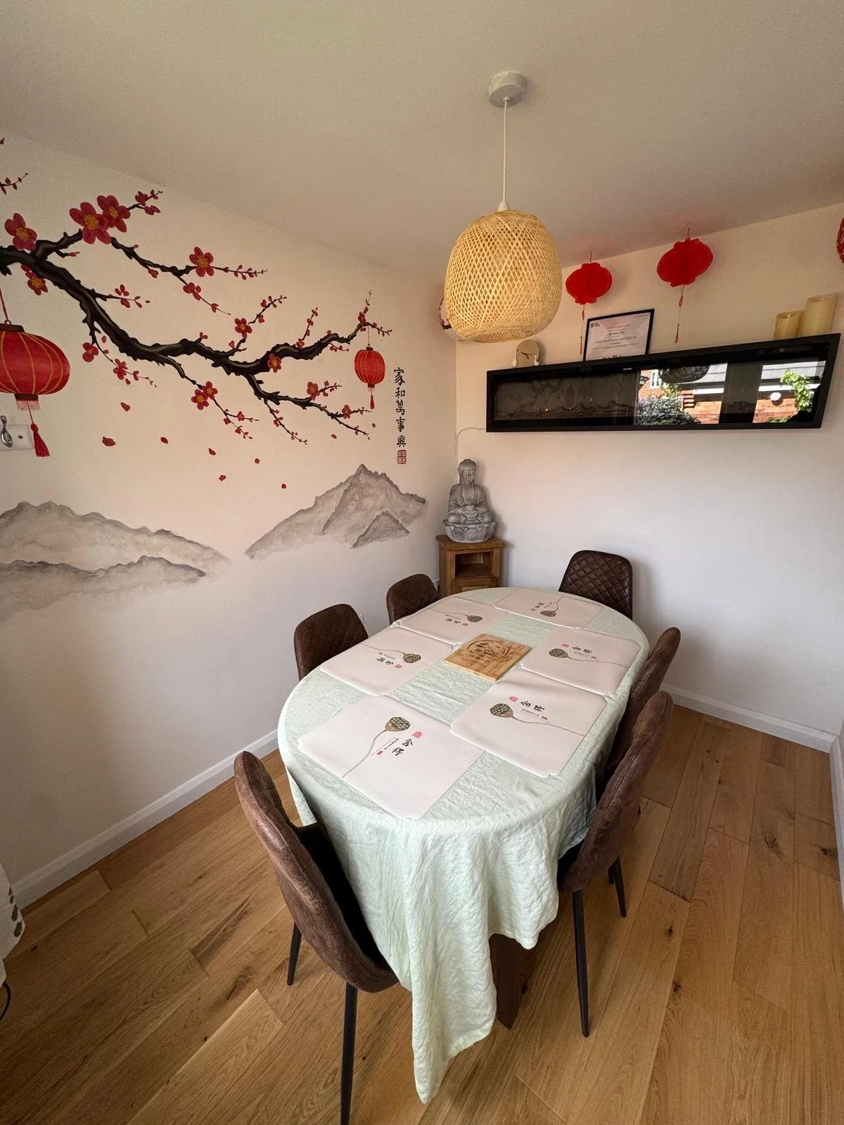 A dining room with a table covered by a green tablecloth and decorated with bamboo-themed placemats, surrounded by six brown upholstered chairs. The wall features a cherry blossom mural with pink flowers and red lanterns, and there is a small Buddha 