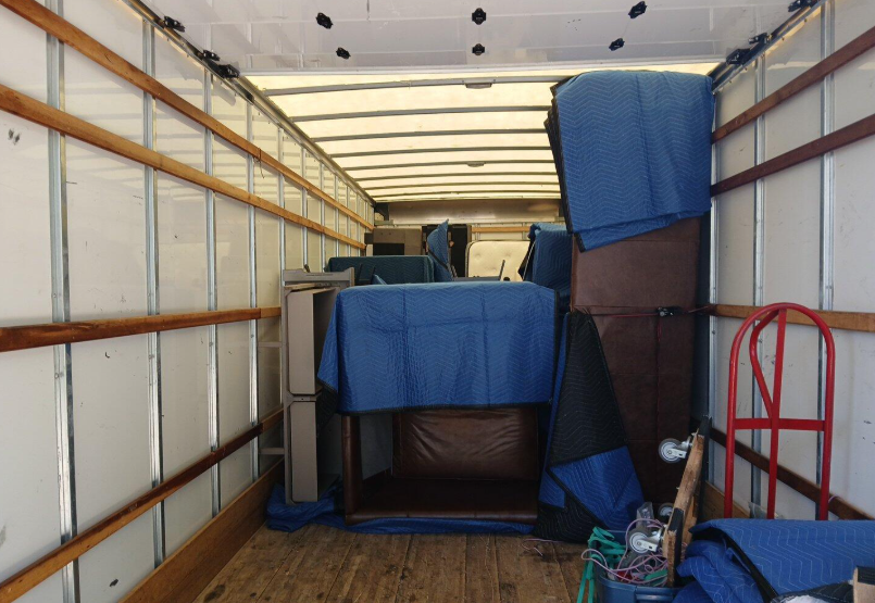 Interior of a moving truck filled with furniture covered in blue moving blankets, stacked and placed for transport.
