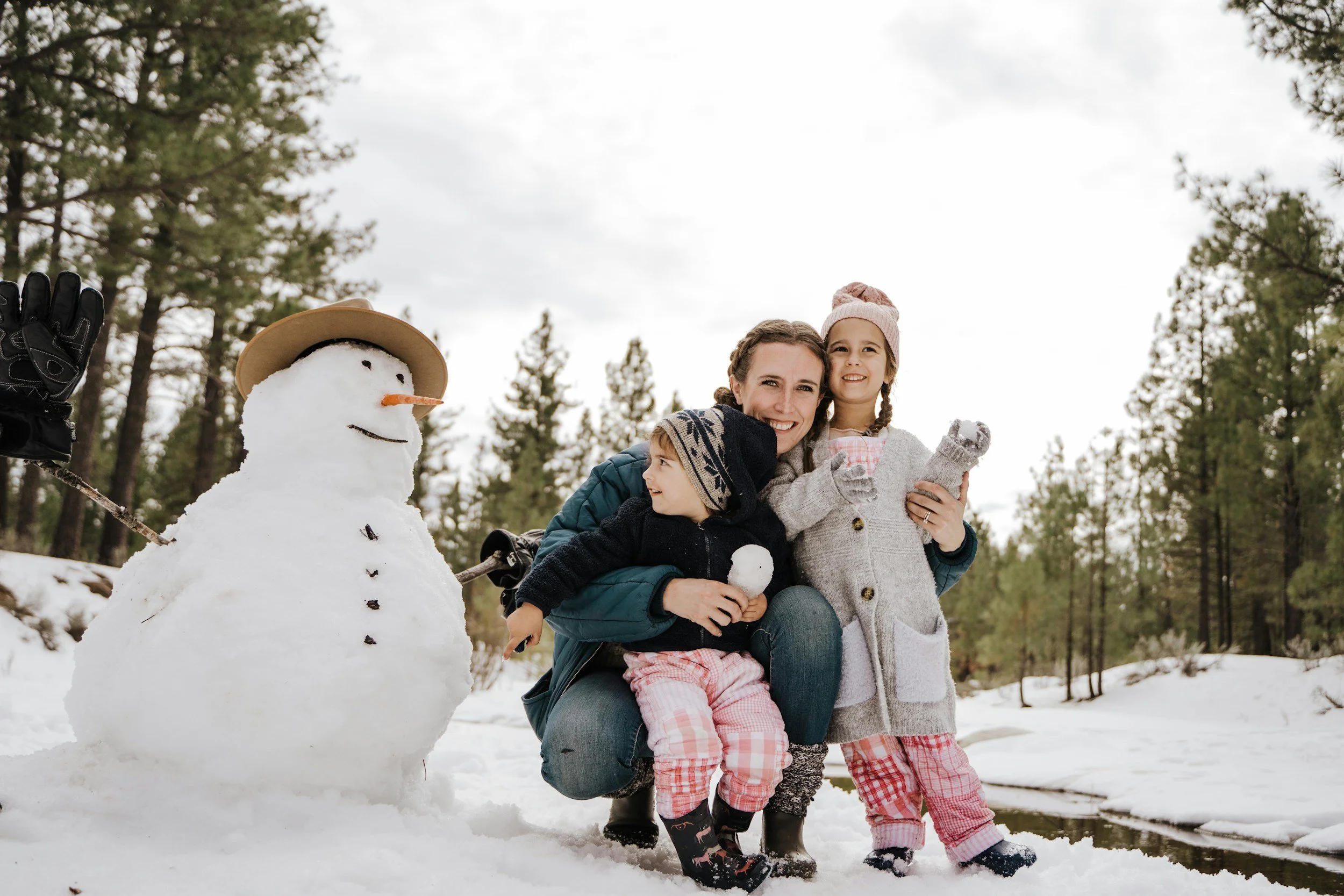 A woman and two children smiling while posing next to a snowman outdoors in a snowy forested area, with cloudy sky above.
