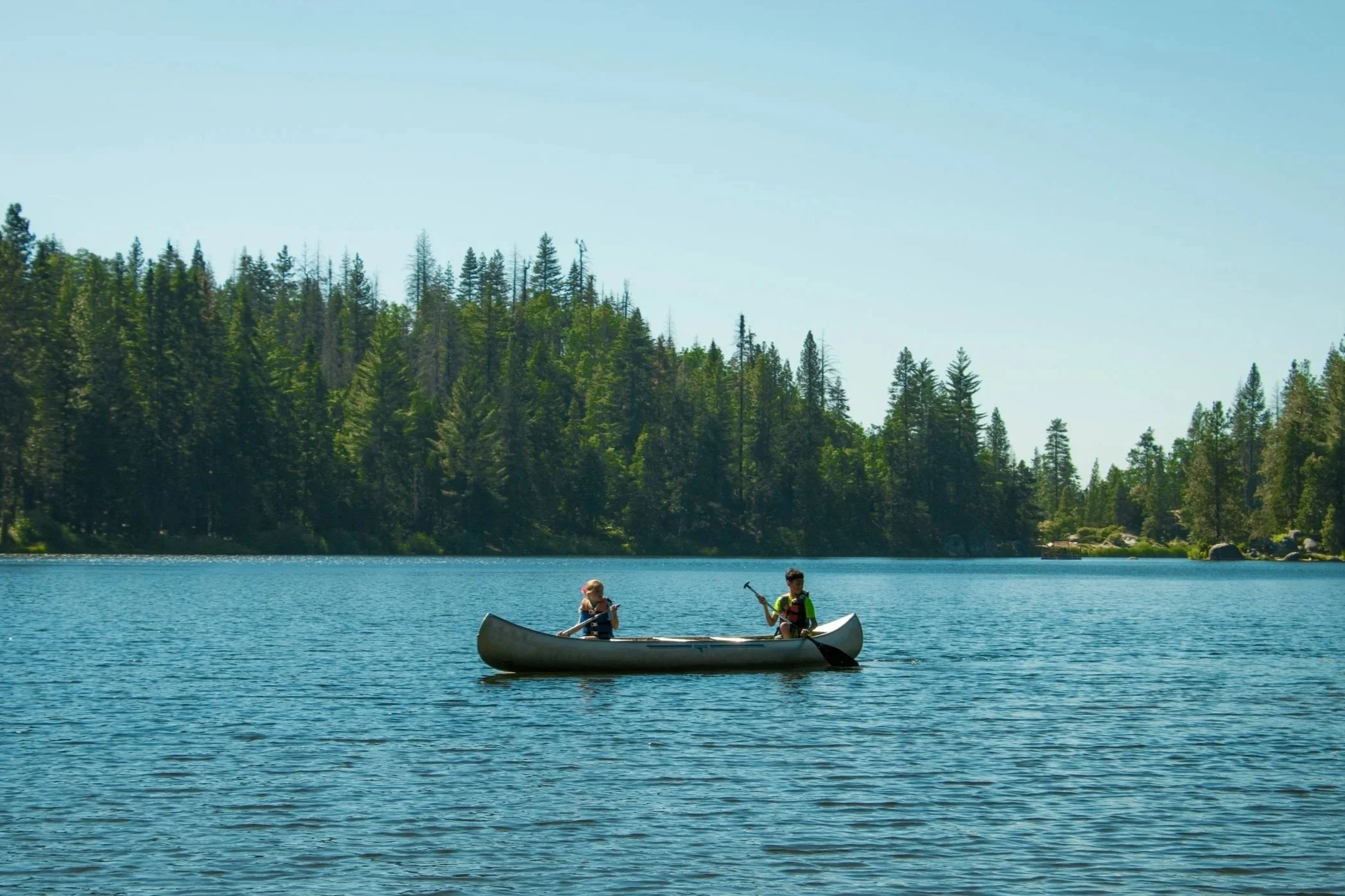 Two children paddleboarding on a lake with a forested hillside in the background during sunny weather.
