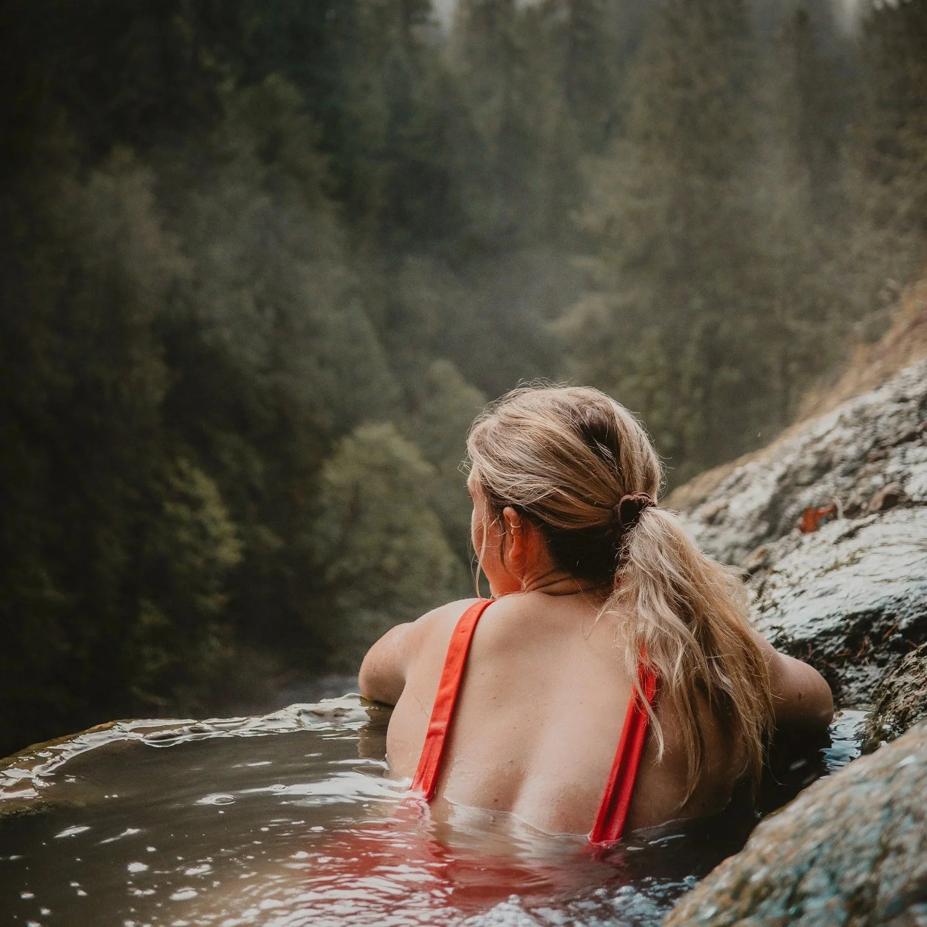 A woman with long blonde hair tied back, wearing an orange tank top, is sitting in a natural water formation, possibly a hot spring or river, surrounded by rocky edges and lush green forest in the background.