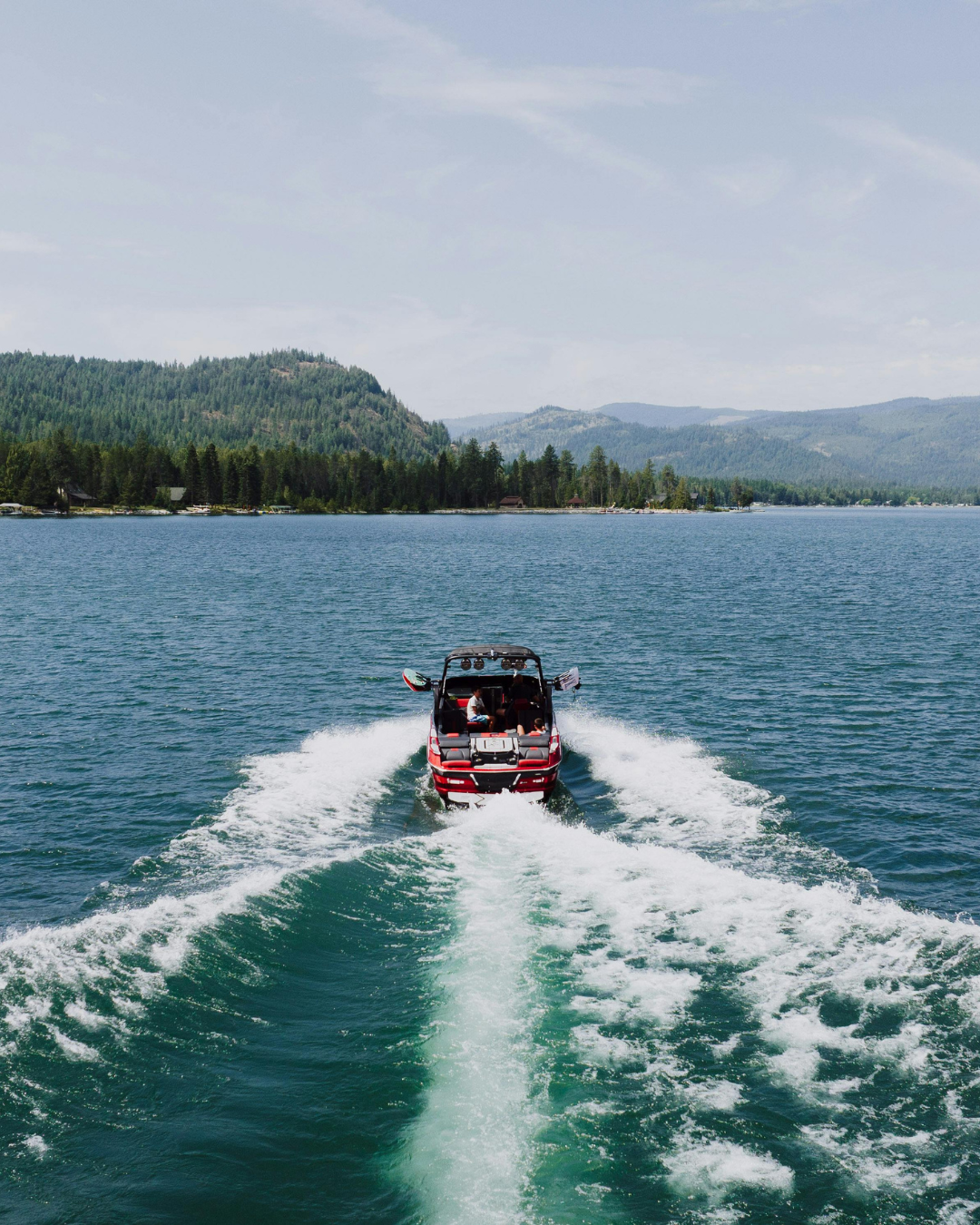 A motorboat speeding across a large body of water, leaving a wake behind. In the background, there are forested hills and mountains under a partly cloudy sky.