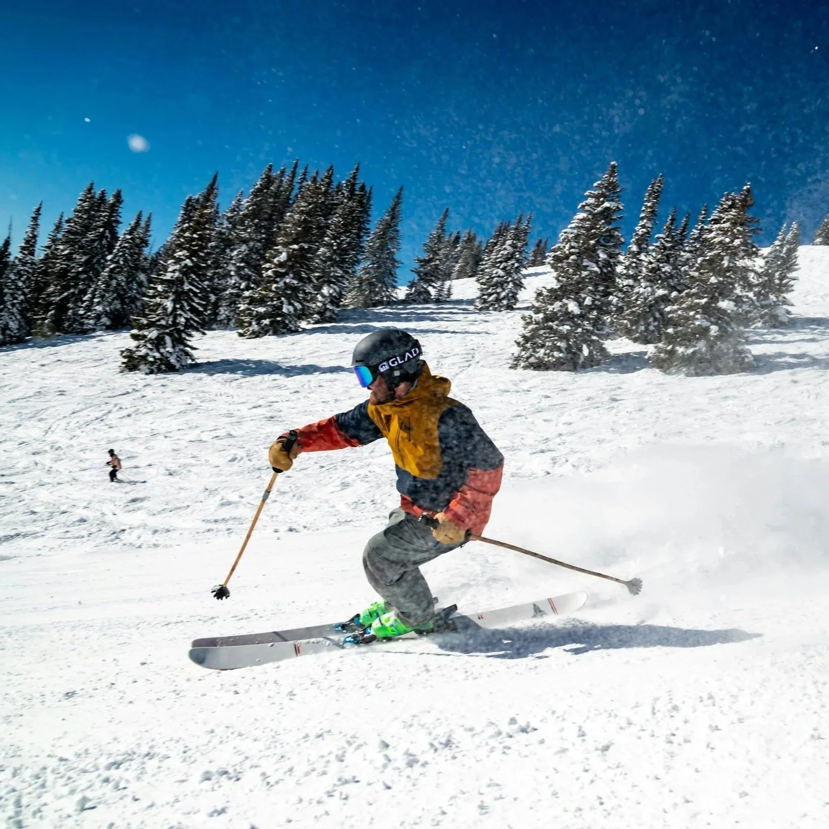 Skier wearing a helmet and yellow jacket skiing on a snowy slope with snow-covered trees and a clear blue sky.