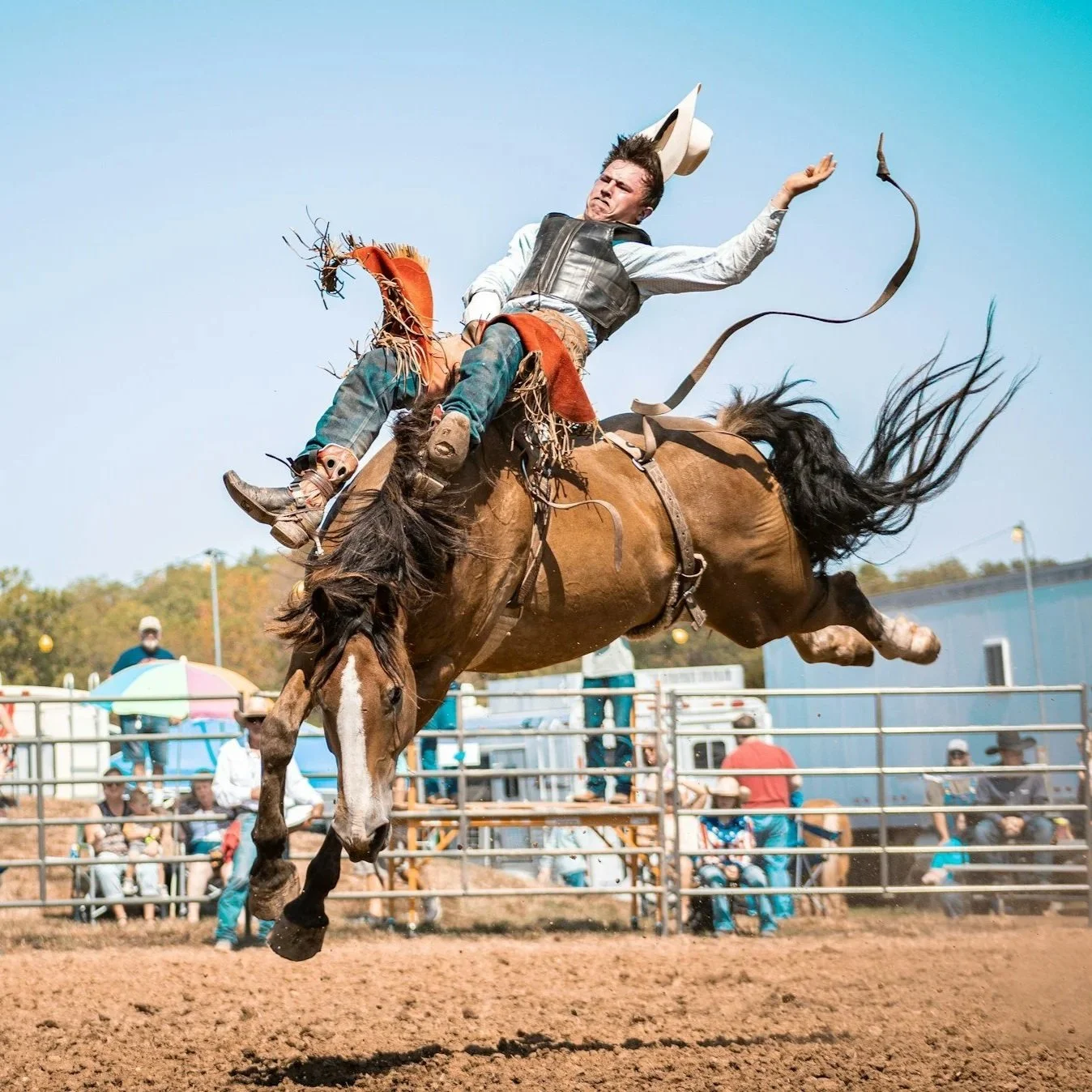 A cowboy is riding a bucking horse at a rodeo event. The cowboy appears to be falling off, with one arm raised and his hat flying off. The background shows spectators and a rodeo arena.