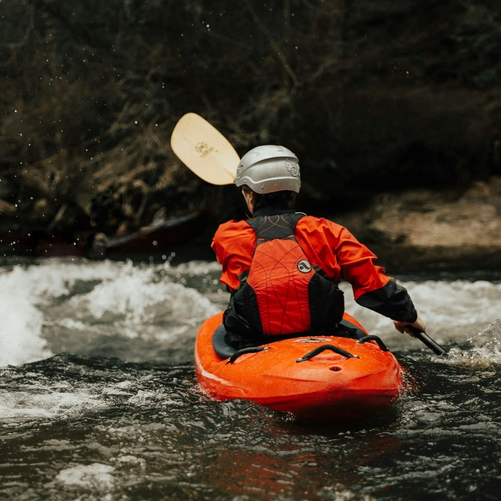 Person in a helmet and orange life vest kayaking through a dark river cave with water splashing around.
