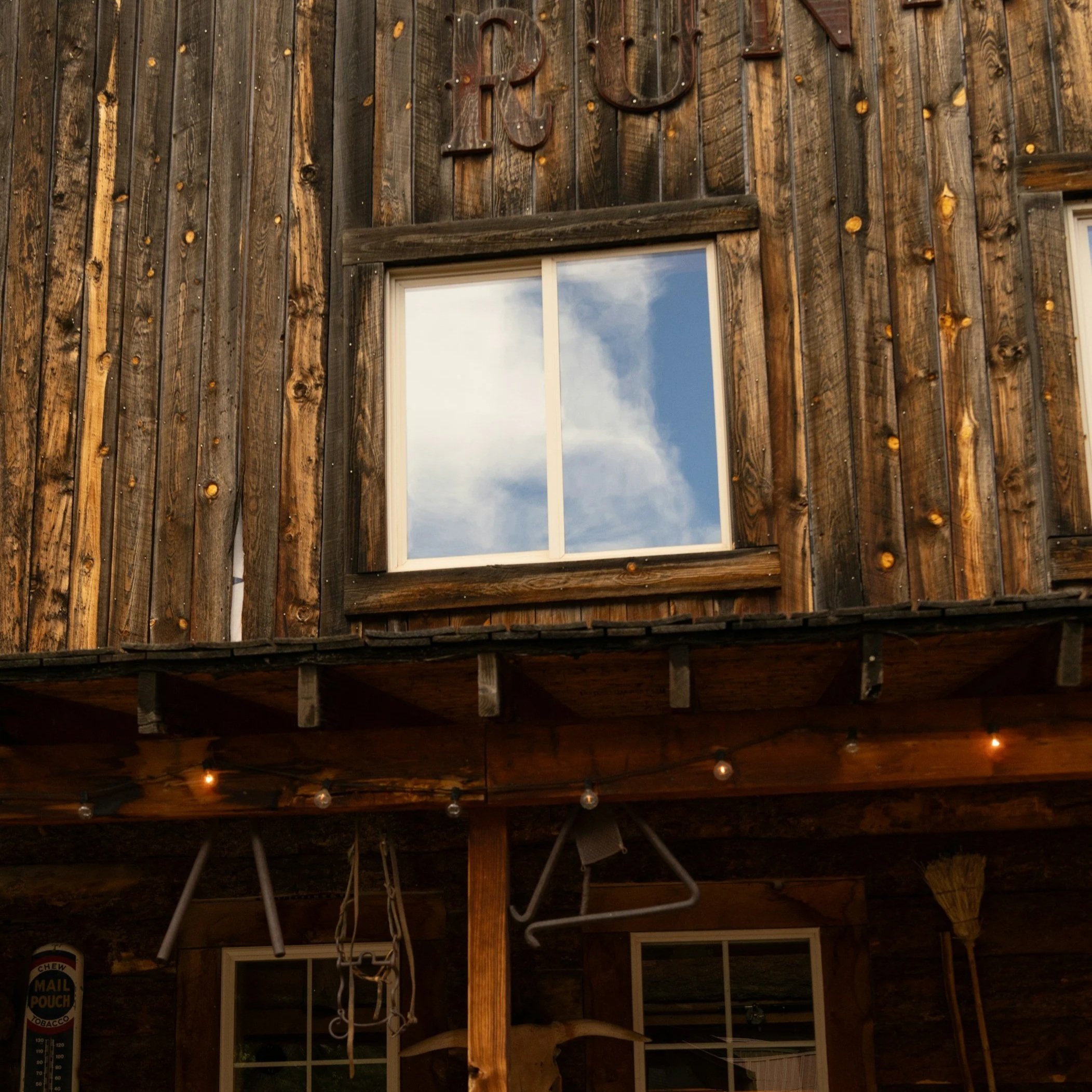 Wooden wall with the large letter 'R' and part of another letter visible, next to a window reflecting the sky, with string lights hanging below.