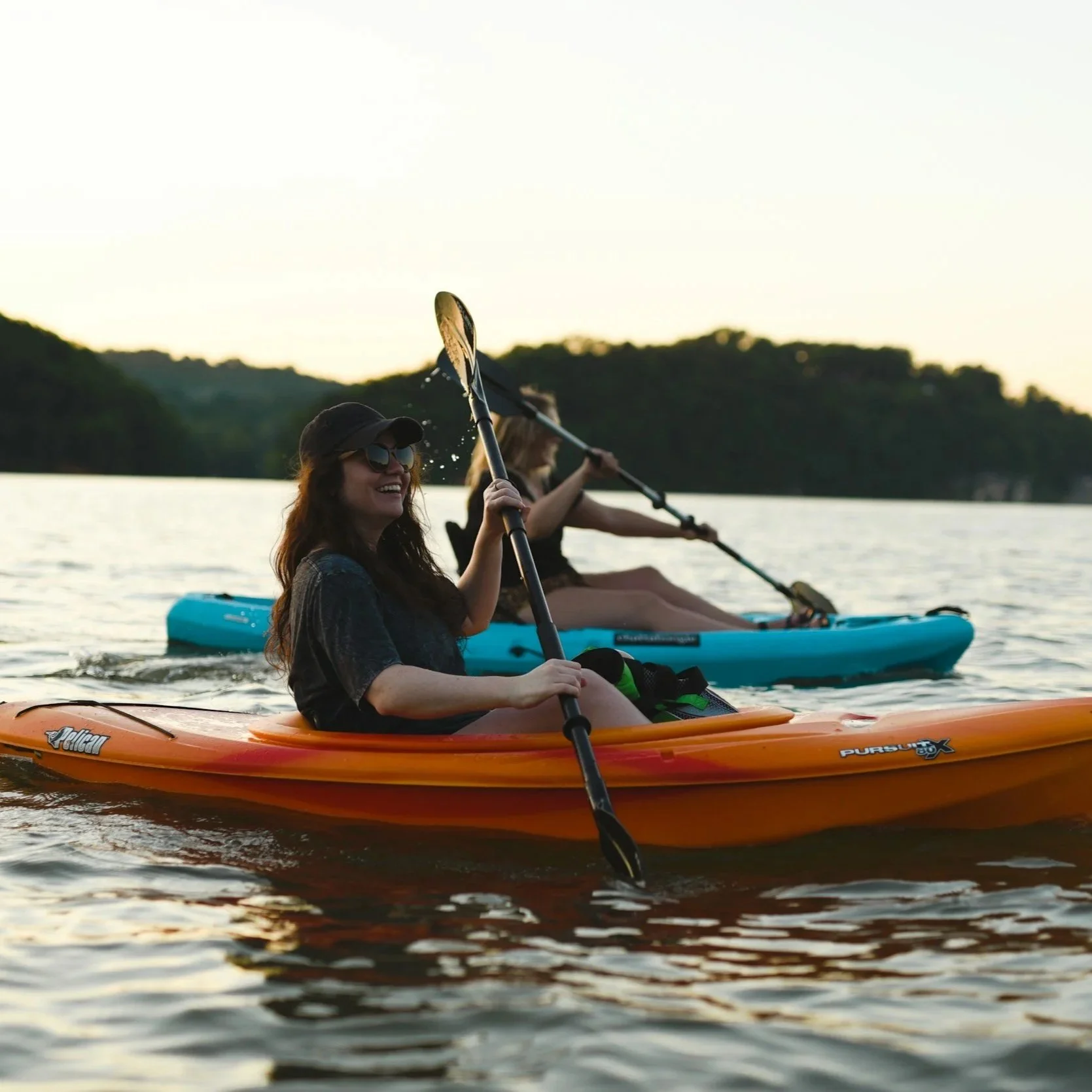 Two women paddleboarding on a lake during sunset, with forested hills in the background.