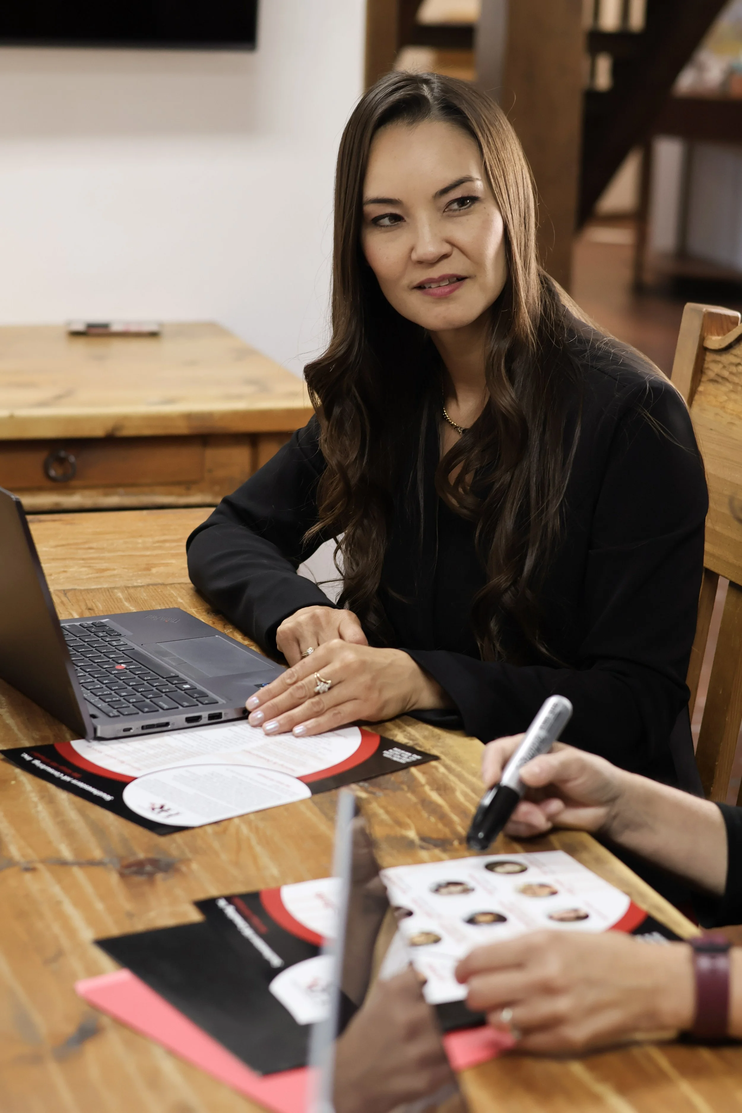 A woman with long brown hair and a black jacket sitting at a wooden table with a laptop, a pen, and some printed menus. She appears to be engaged in conversation or thought, with friends or colleagues.
