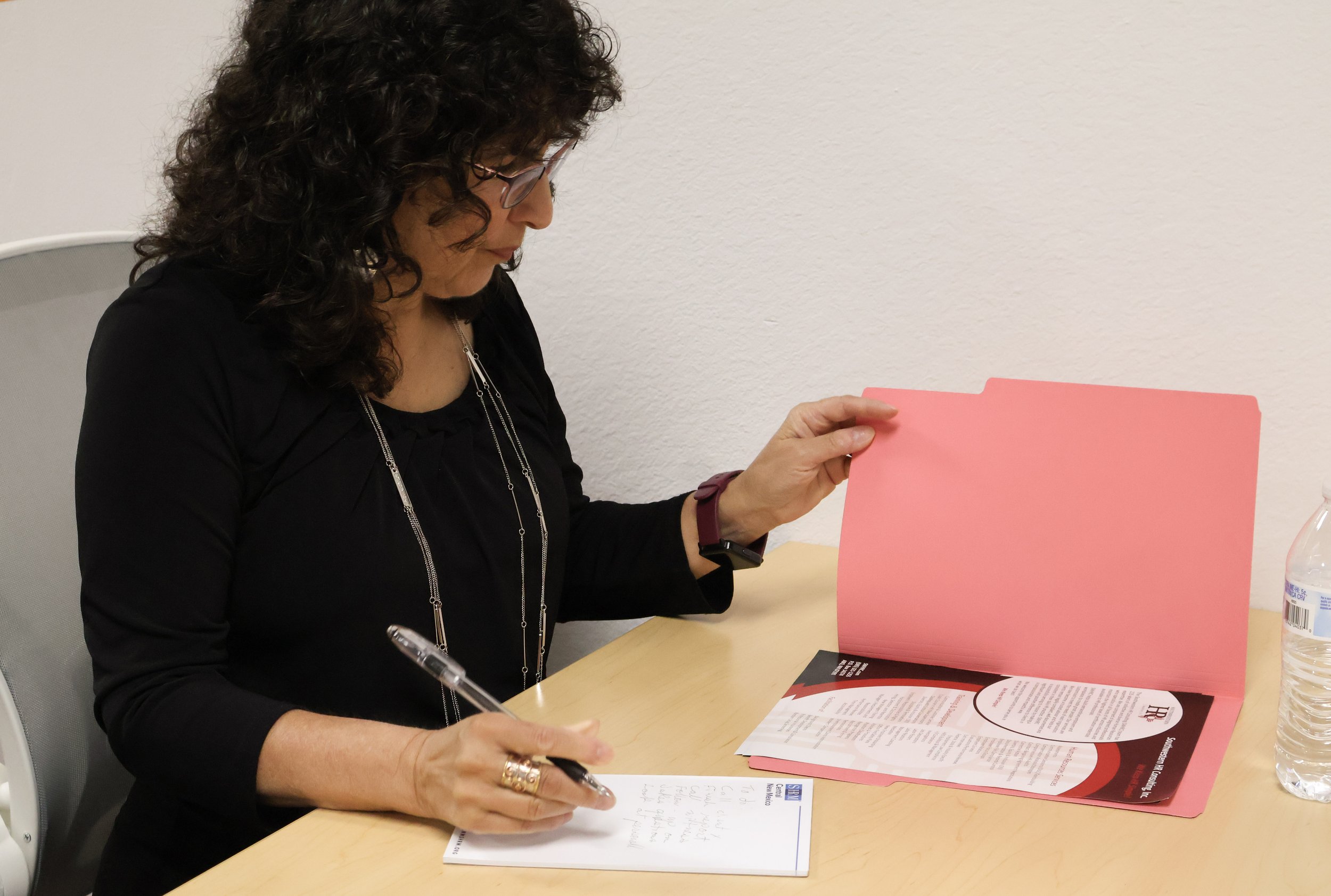 A woman with curly dark hair and glasses sitting at a desk, reading a pink folder while taking notes with a pen.