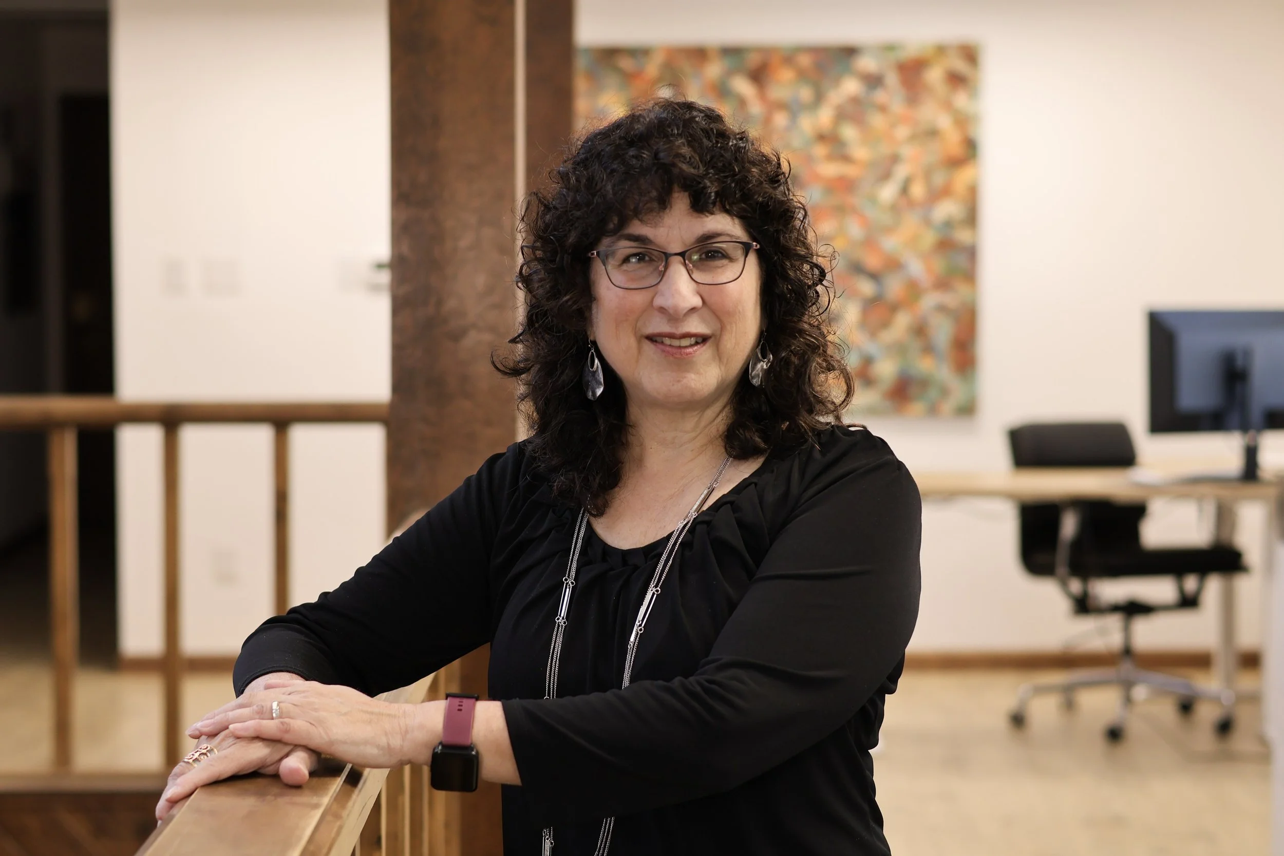 A woman with curly dark hair and glasses smiling, sitting at a wooden table in an office environment, with a colorful abstract painting on the wall behind her.