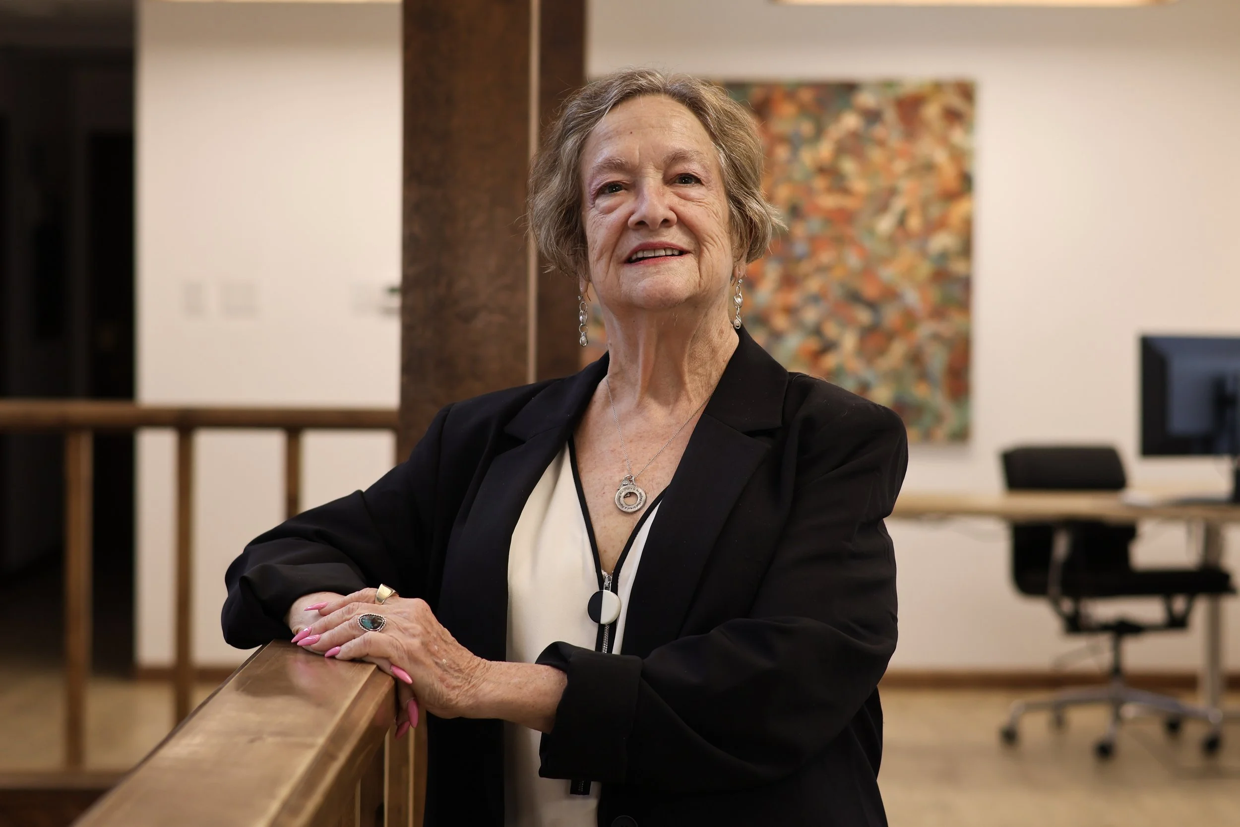 An elderly woman with short gray hair and light skin sitting at a wooden table with her arms crossed, wearing a black blazer, pearl earrings, a pendant necklace, and pink nail polish, in an indoor setting with artwork on the wall behind her.