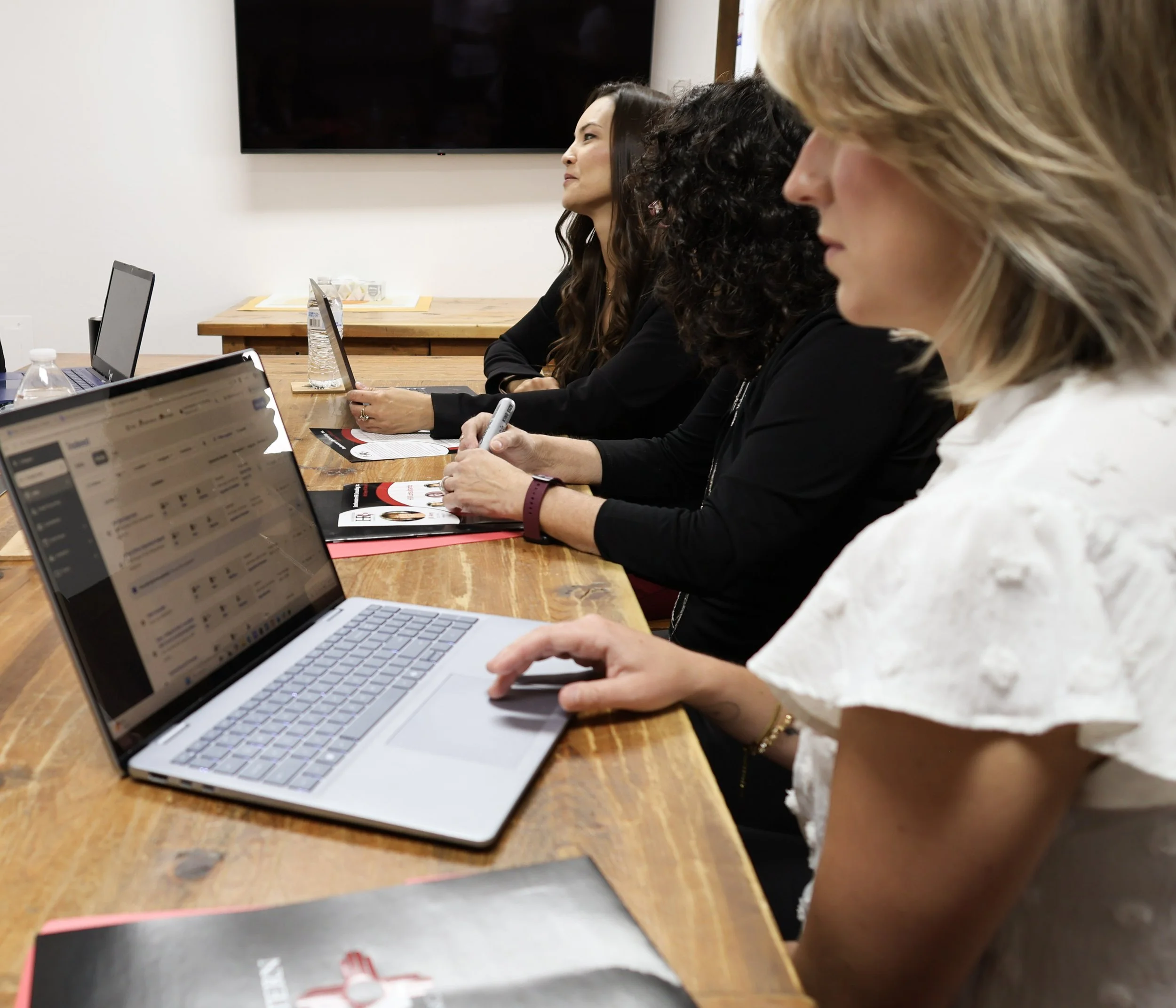 Three women sitting at a wooden table during a meeting or discussion. Laptops, notebooks, and water bottles are on the table. One woman is holding a pen, and another has a brochure or magazine in front of her. They are in a conference room with a large black screen or TV on the wall.