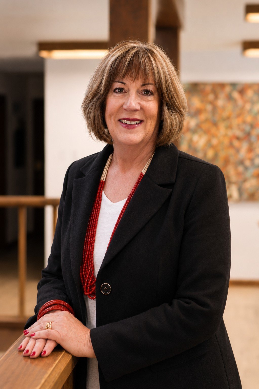 A woman with shoulder-length brown hair and a warm smile, wearing a black blazer, white top, and red beaded jewelry, posing indoors against a background with soft lighting and a colorful artwork.