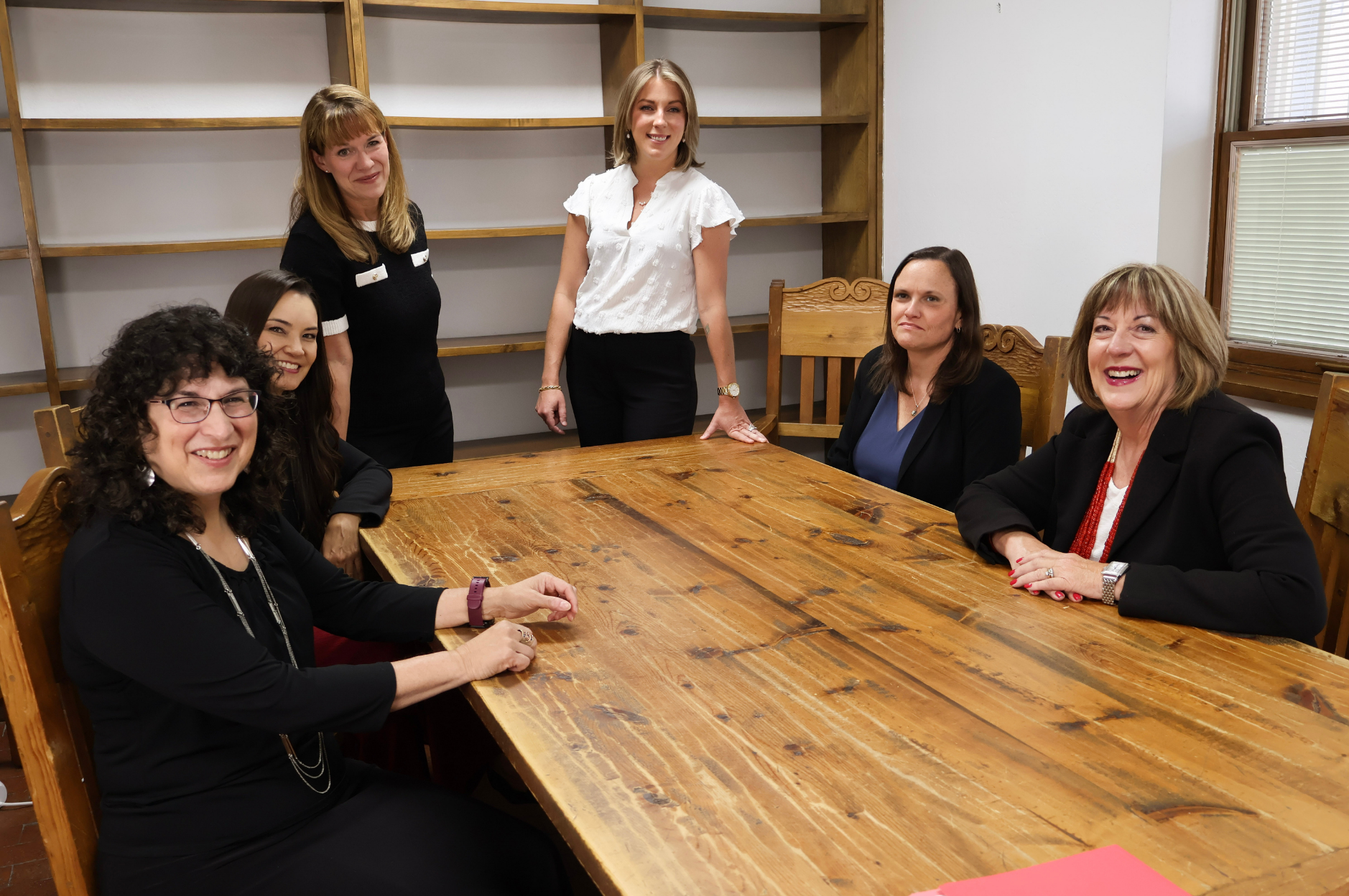 Six women are gathered around a large wooden conference table in a room with wooden shelves and windows with blinds. Four women are seated, and two are standing behind them, all smiling or with neutral expressions.