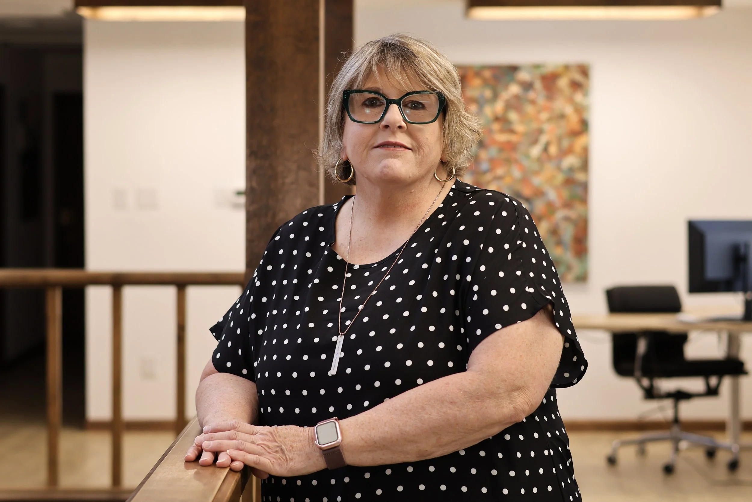 A middle-aged woman with short blonde hair and black-rimmed glasses stands indoors. She is wearing a black dress with white polka dots, silver hoop earrings, a necklace, and a smartwatch. She is leaning on a wooden railing with her arms crossed. Behind her, there is an abstract colorful painting on the wall, a wooden post, and some tables and chairs.