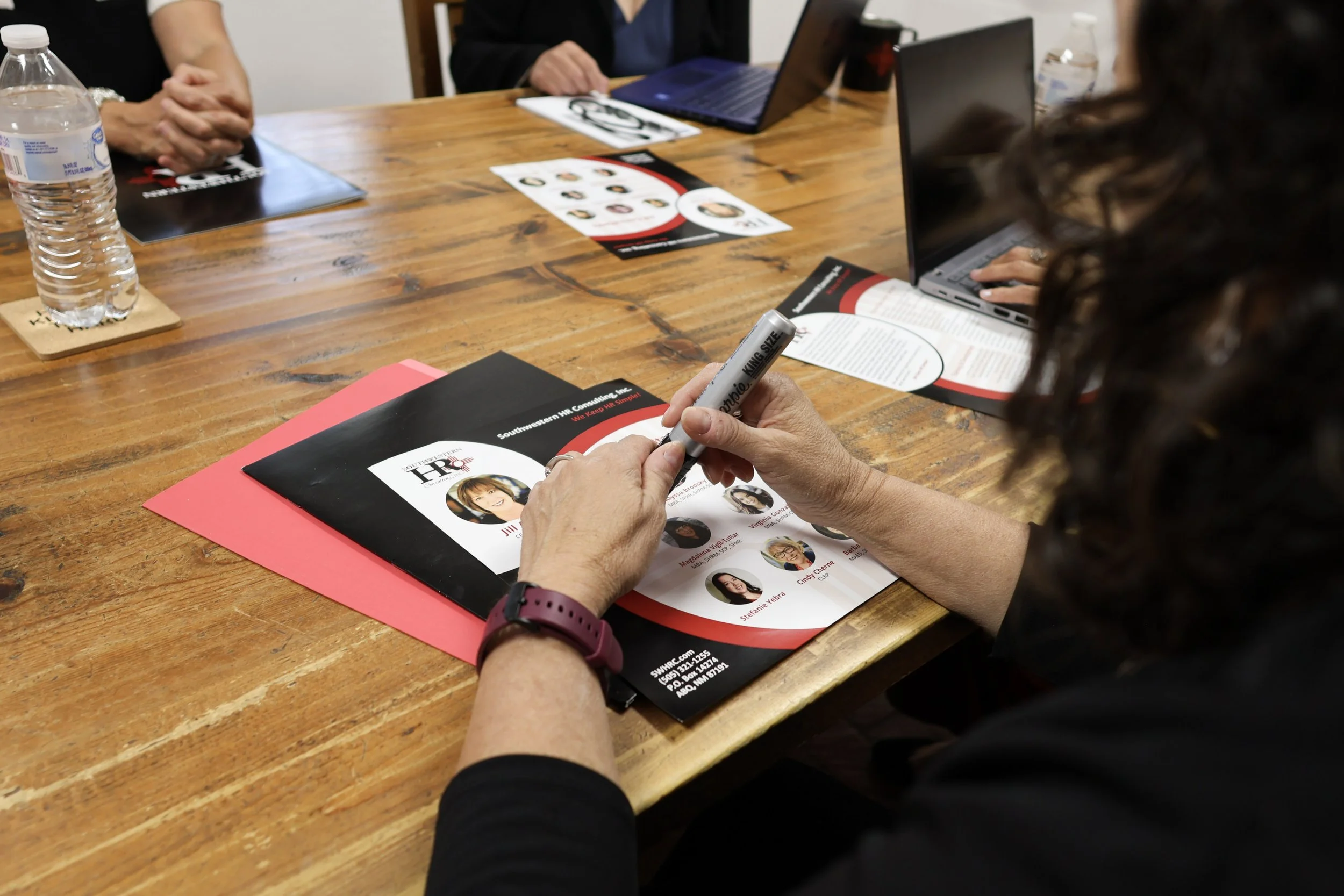 People sitting around a wooden table with laptops, pamphlets, and a water bottle. One person is writing on a pamphlet while others are sitting with hands clasped or resting.