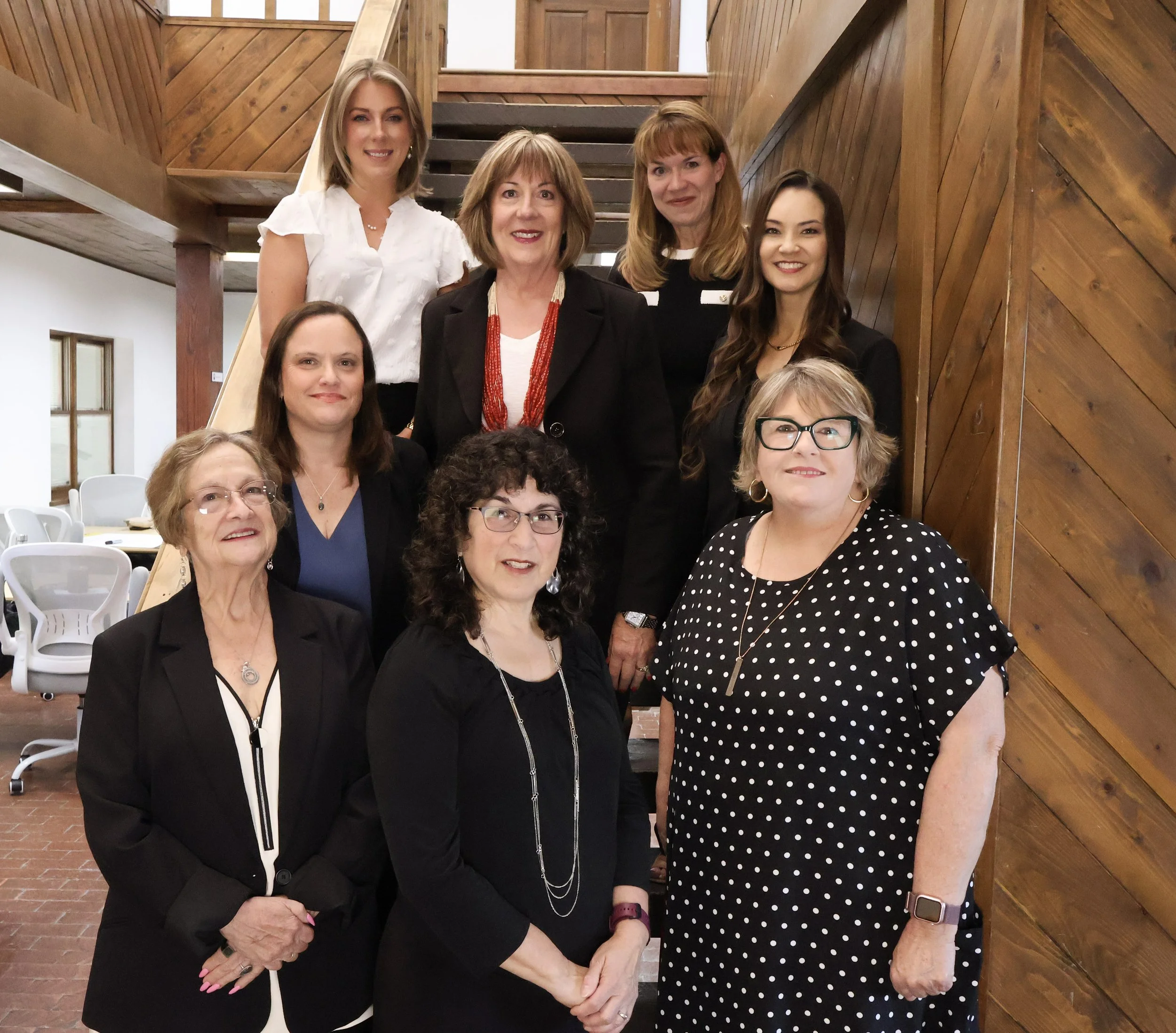Group of women posing on a staircase inside a building with wooden walls, dressed in formal and business attire.