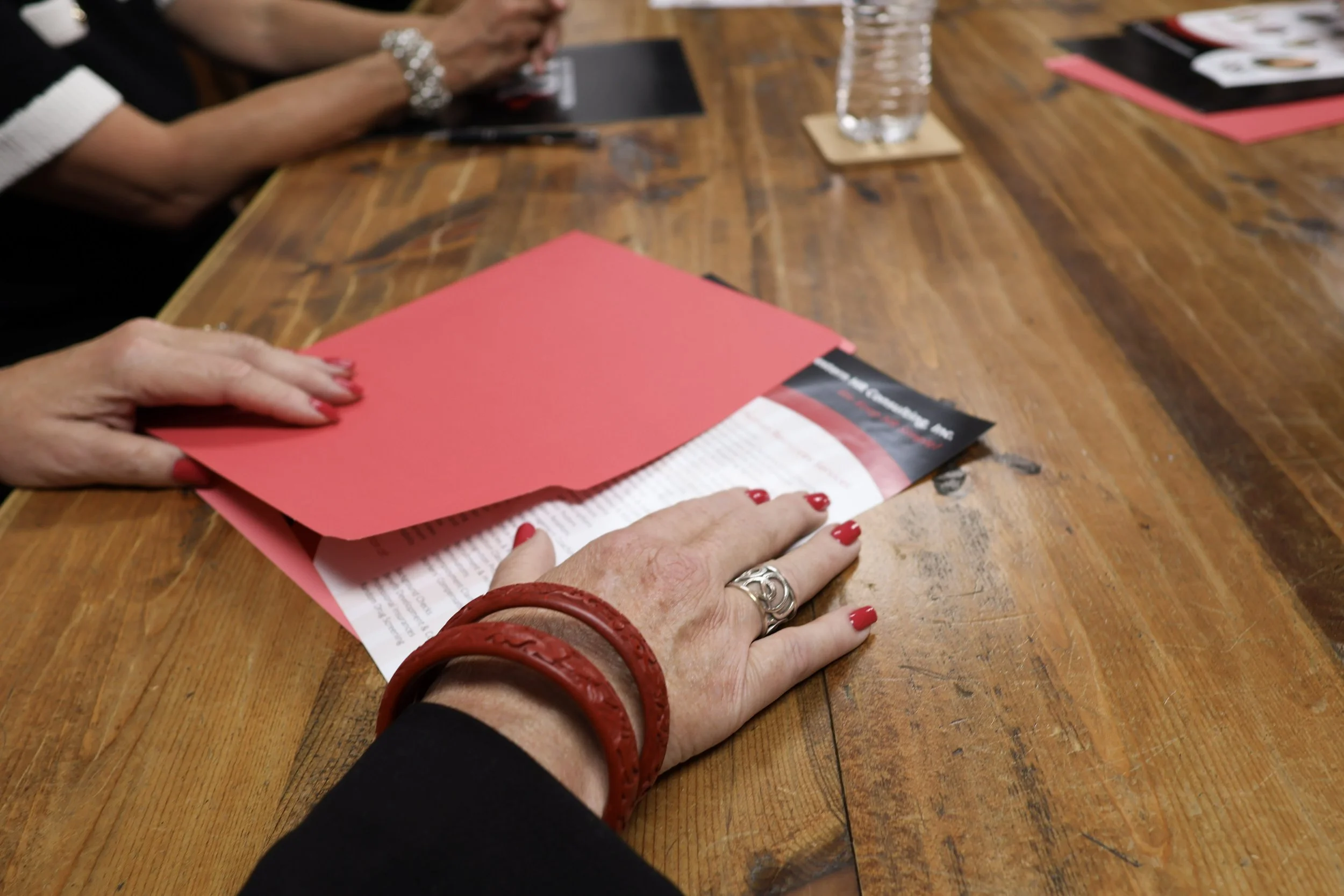 Close-up of a woman's hands with red painted nails, wearing silver jewelry and a red bracelet, resting on a wooden table, with pink folders and a glass of water nearby.