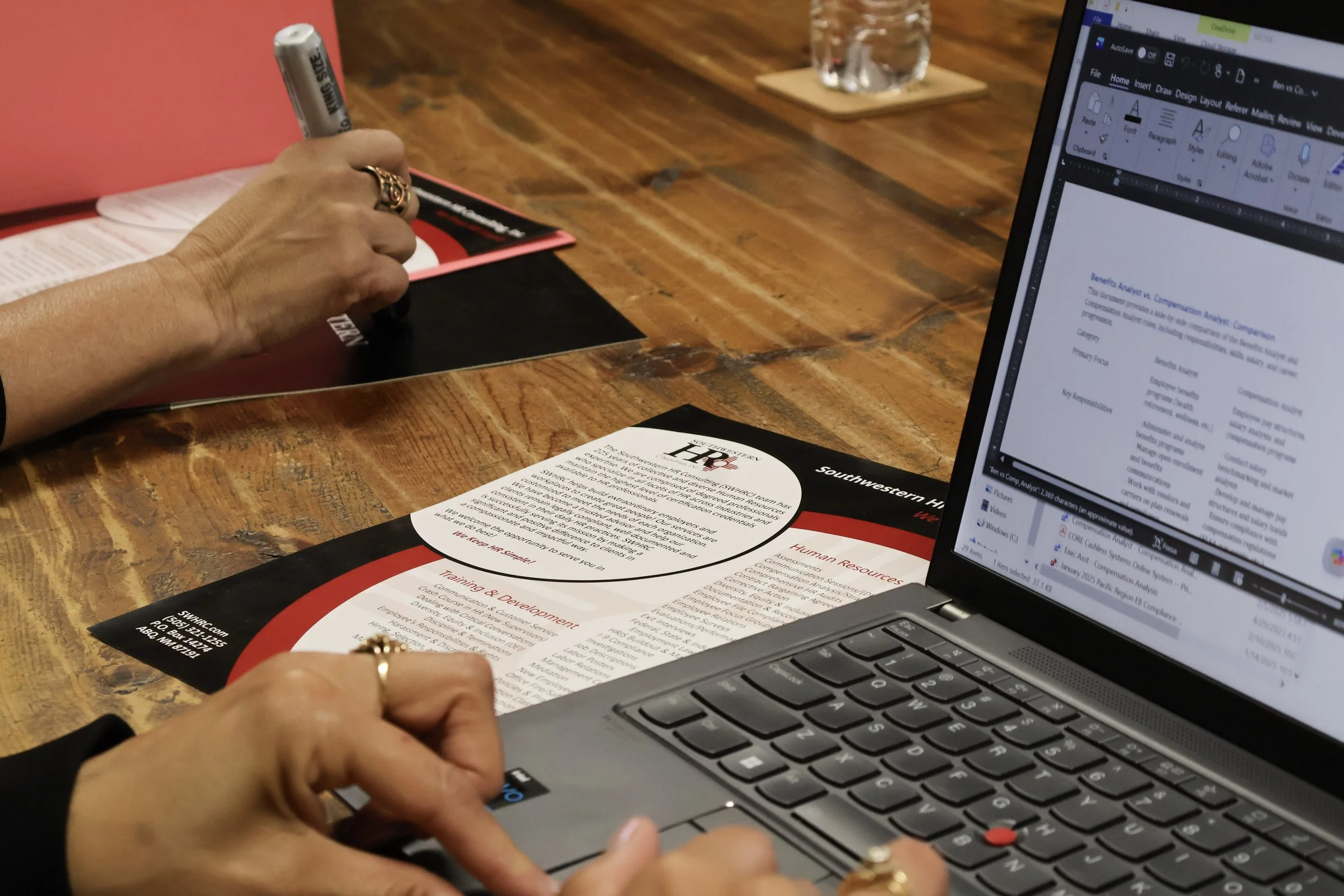 Person holding pen and paper while sitting at a wooden table with a laptop and a brochure for Human Resources resources.