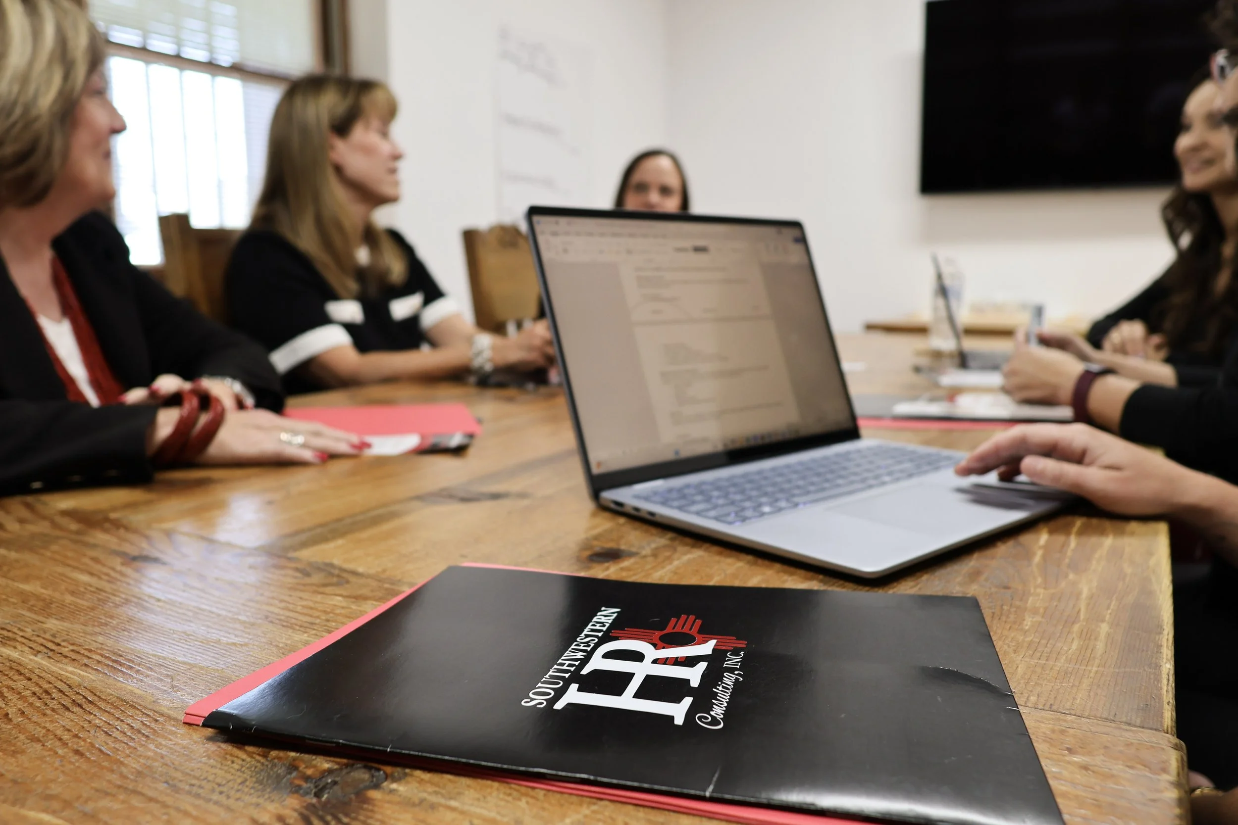 A group of women sitting around a wooden table in a conference room. There are laptops, notebooks, and a folder on the table. The room has windows with blinds and a large screen on the wall.