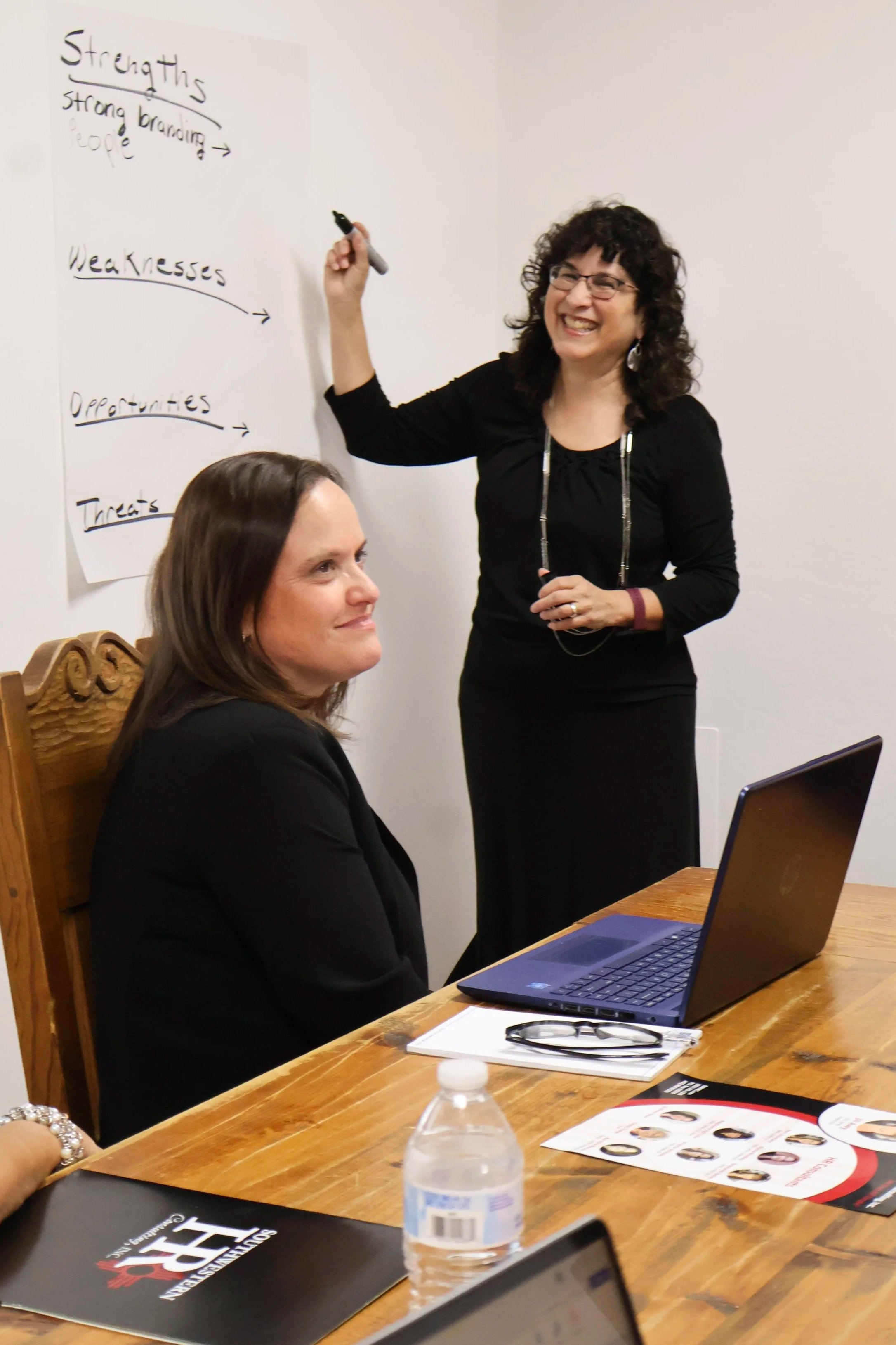 A woman with curly hair and glasses is standing and smiling while writing on a whiteboard with handwritten notes. Seated at a wooden table is another woman with straight hair working on a laptop. The table has a water bottle, a pamphlet with photos, a notebook, and other items.