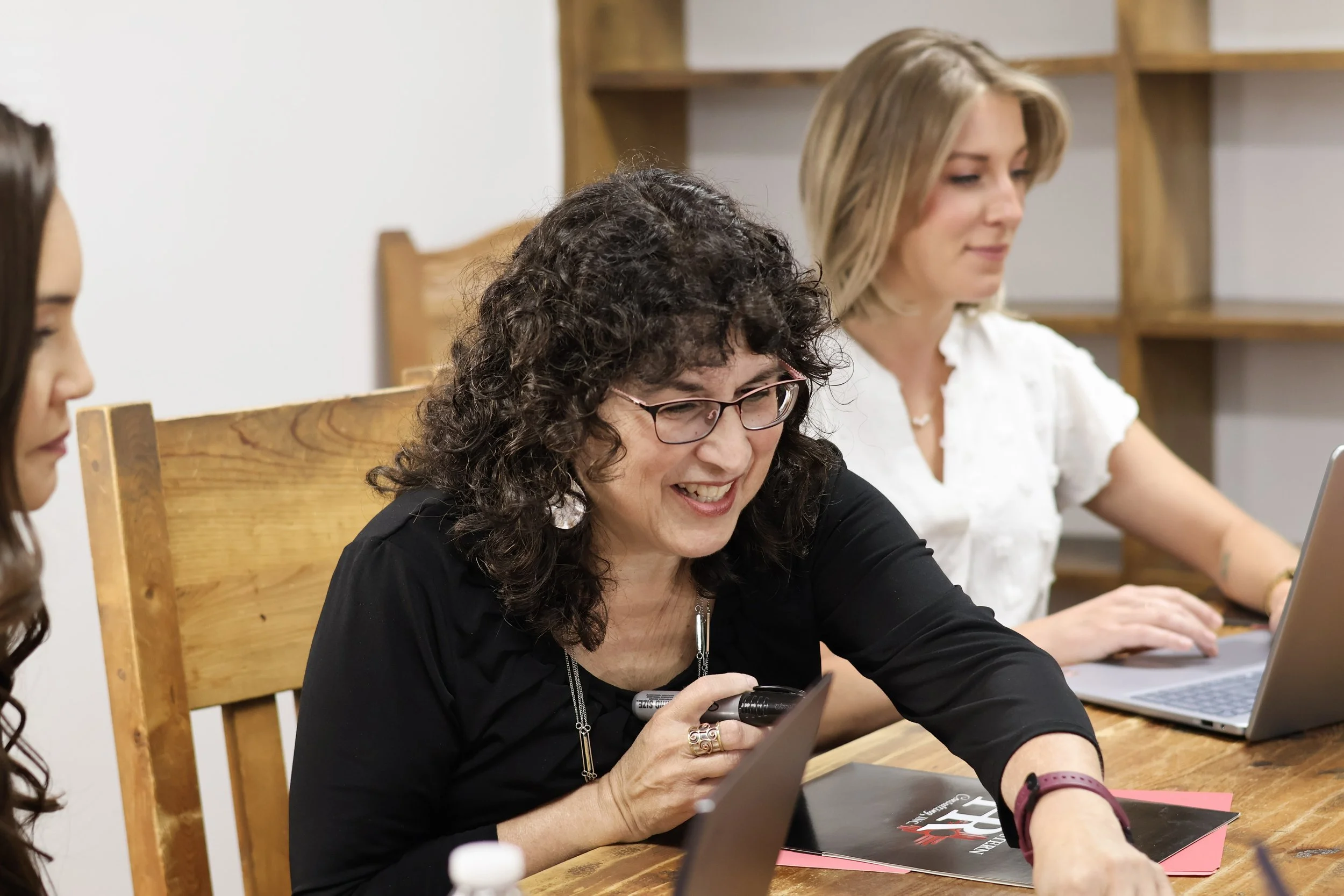 Three women sitting at a wooden table working with laptops and notebooks, smiling and engaged in their tasks.