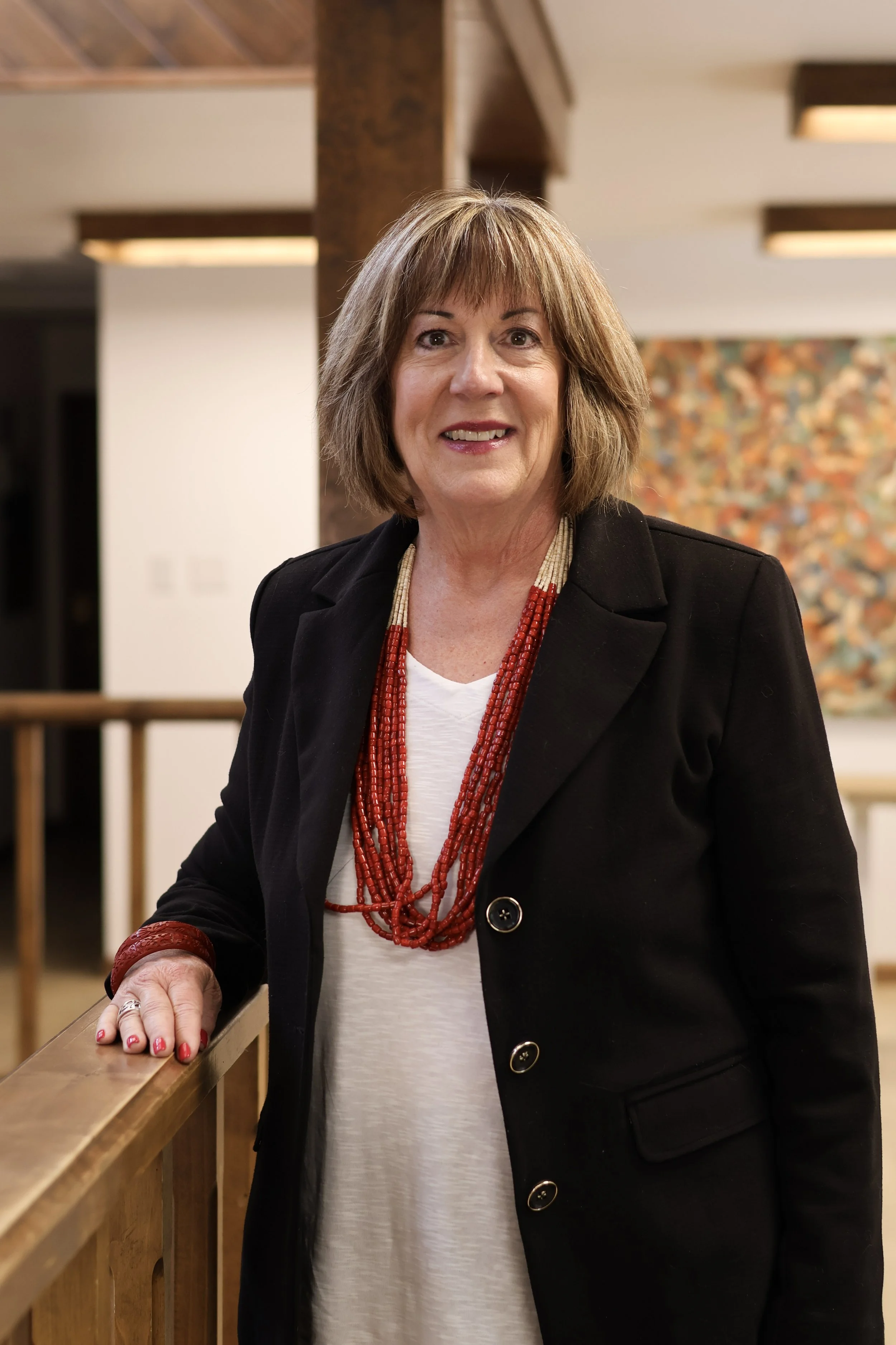 A woman with shoulder-length brown hair, wearing a black blazer with gold buttons, a white top, and red beaded necklaces, leaning on a wooden railing inside a building with warm lighting.