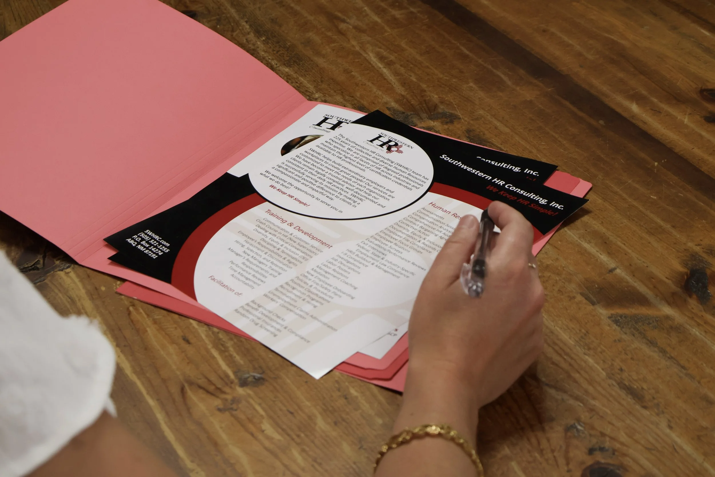 A person writing on a printed flyer at a wooden table with a pink folder nearby.