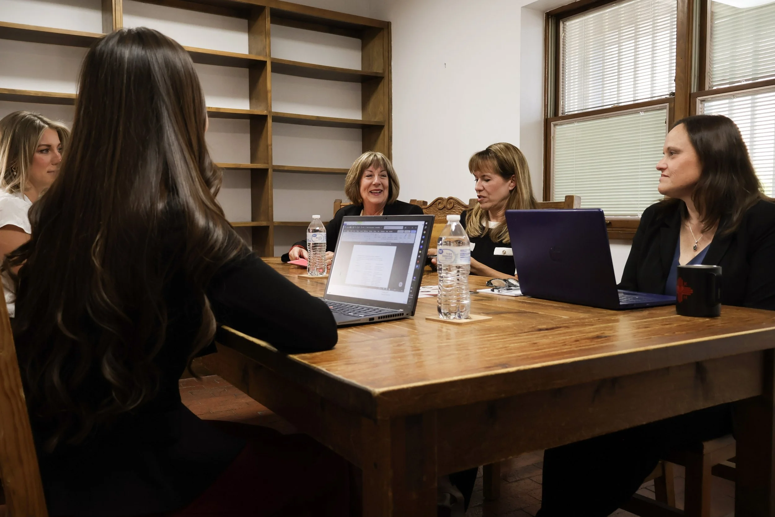 Group of women sitting around a large wooden table in a meeting room, engaged in conversation, with laptops and water bottles on the table.