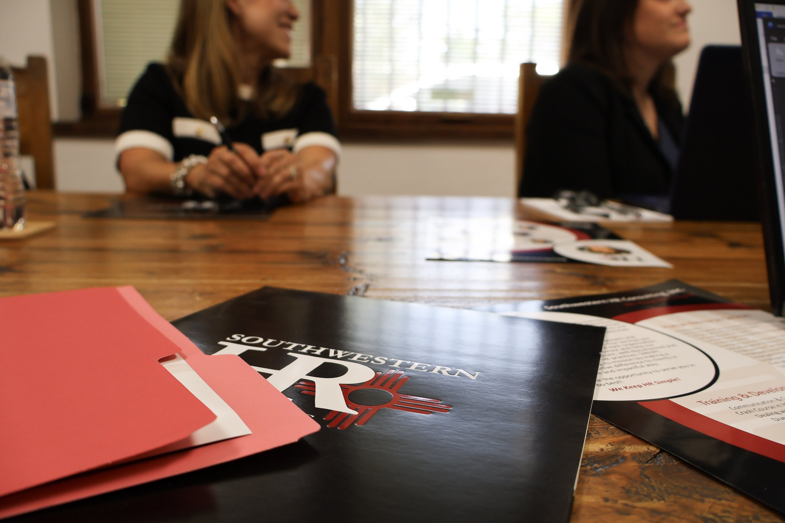 Blurred image of two women sitting at a wooden table in a room with large windows and blinds, engaging in conversation. On the table are various printed materials, including a black and red pamphlet, a brochure, and a pink folder.