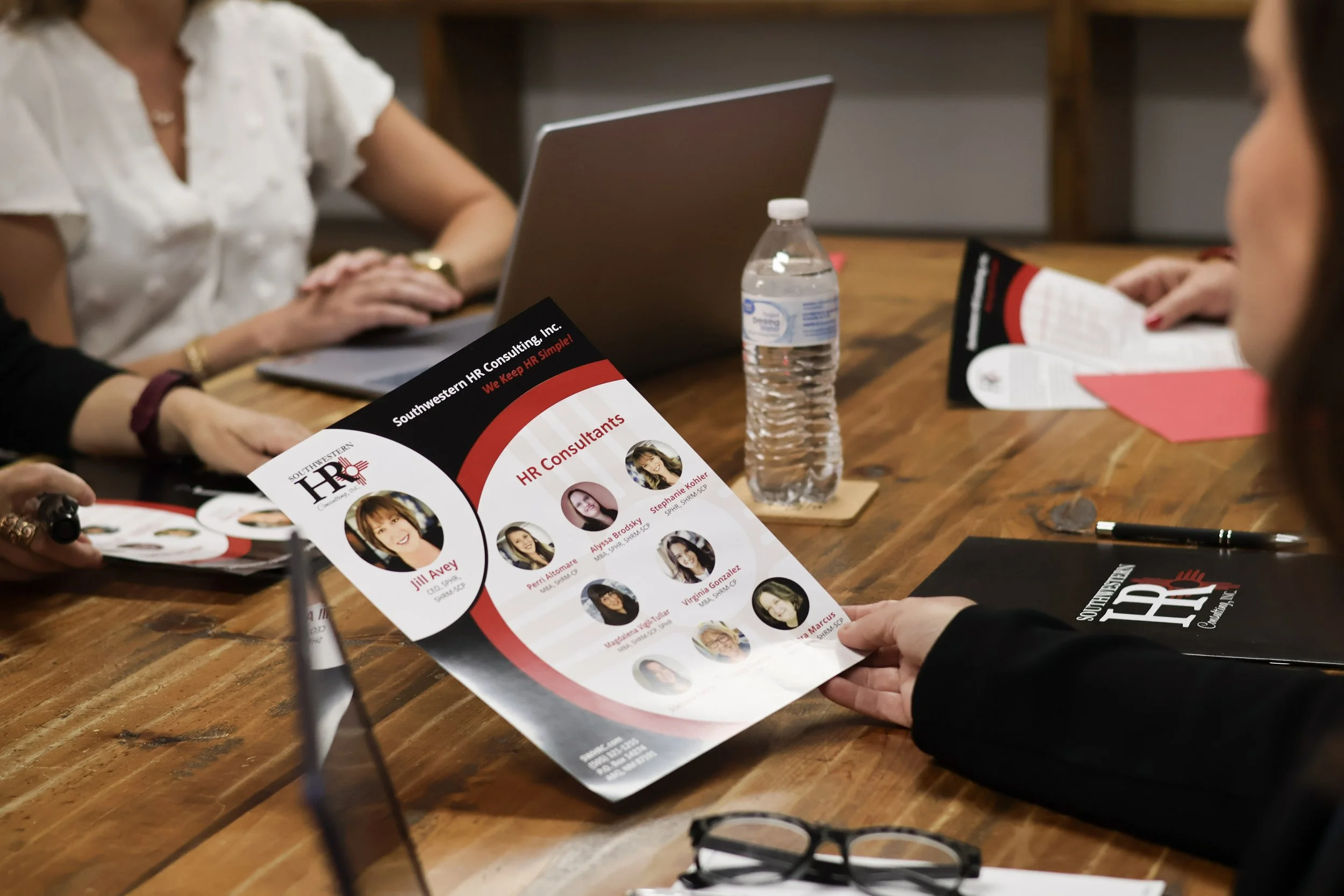 People at a wooden table looking at a brochure featuring HR consultants, with laptops, a water bottle, and pamphlets on the table.