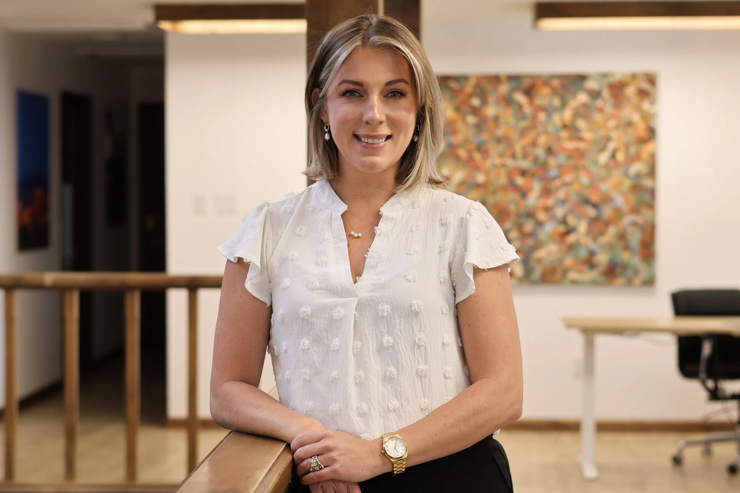 A woman with shoulder-length blond hair, wearing earrings, a pearl necklace, a gold watch, and a white blouse with textured polka dots, standing in an art gallery. She is leaning on a wooden railing with her right arm, smiling at the camera. The background features a large, colorful abstract painting on the wall.