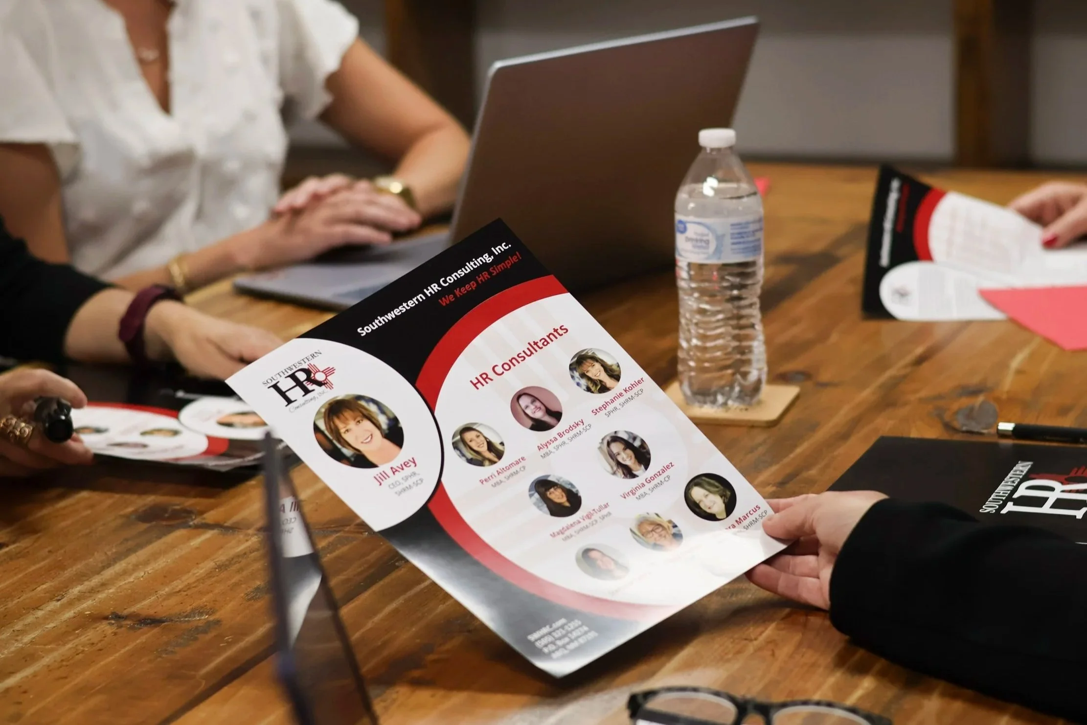 People sitting around a wooden table, holding and discussing a brochure for HR consulting services featuring photos of various women professionals.