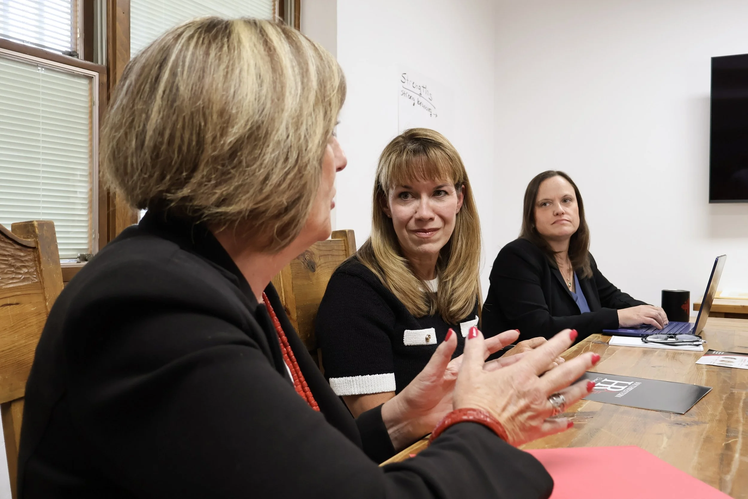 Three women engaged in a discussion around a wooden table in a bright room, with laptop and printed materials in front of them.