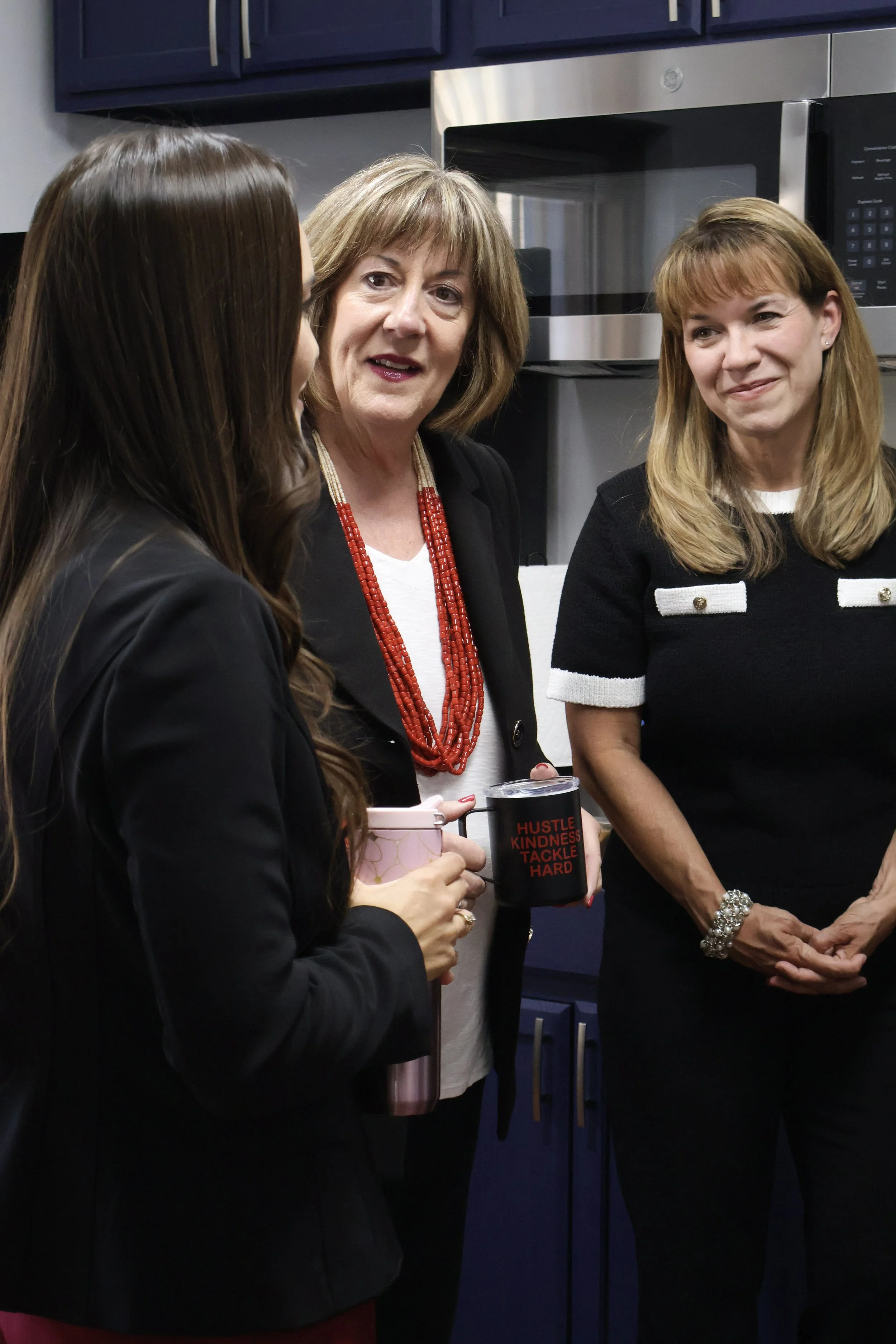 Three women standing in a kitchen, engaged in conversation, with one holding a pink mug and another holding a black mug that reads, 'HUSTLE KINDNESS TACKLE HARD.'