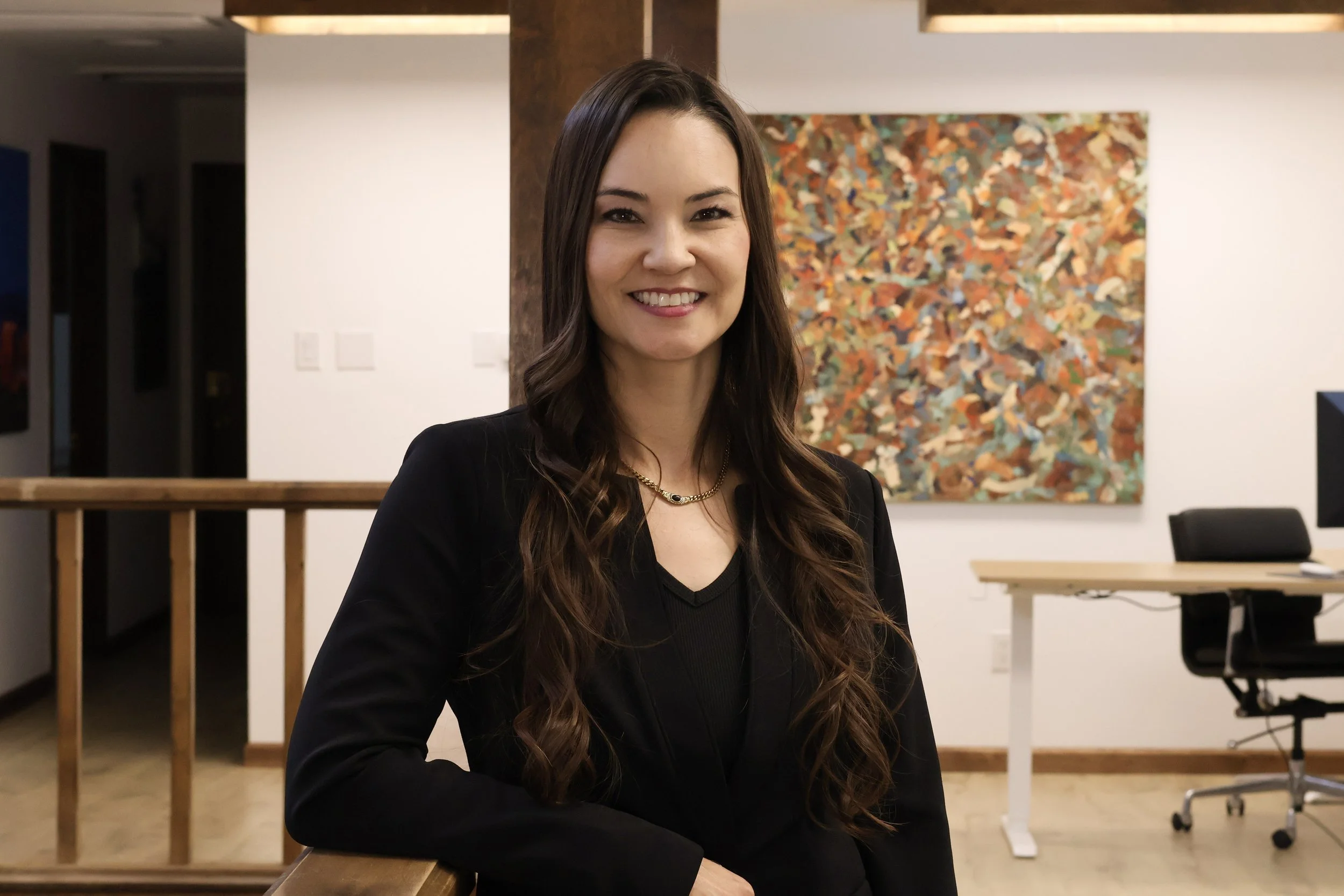 A woman with long brown hair smiling at the camera in an art gallery or office space, with abstract artwork on the wall behind her.