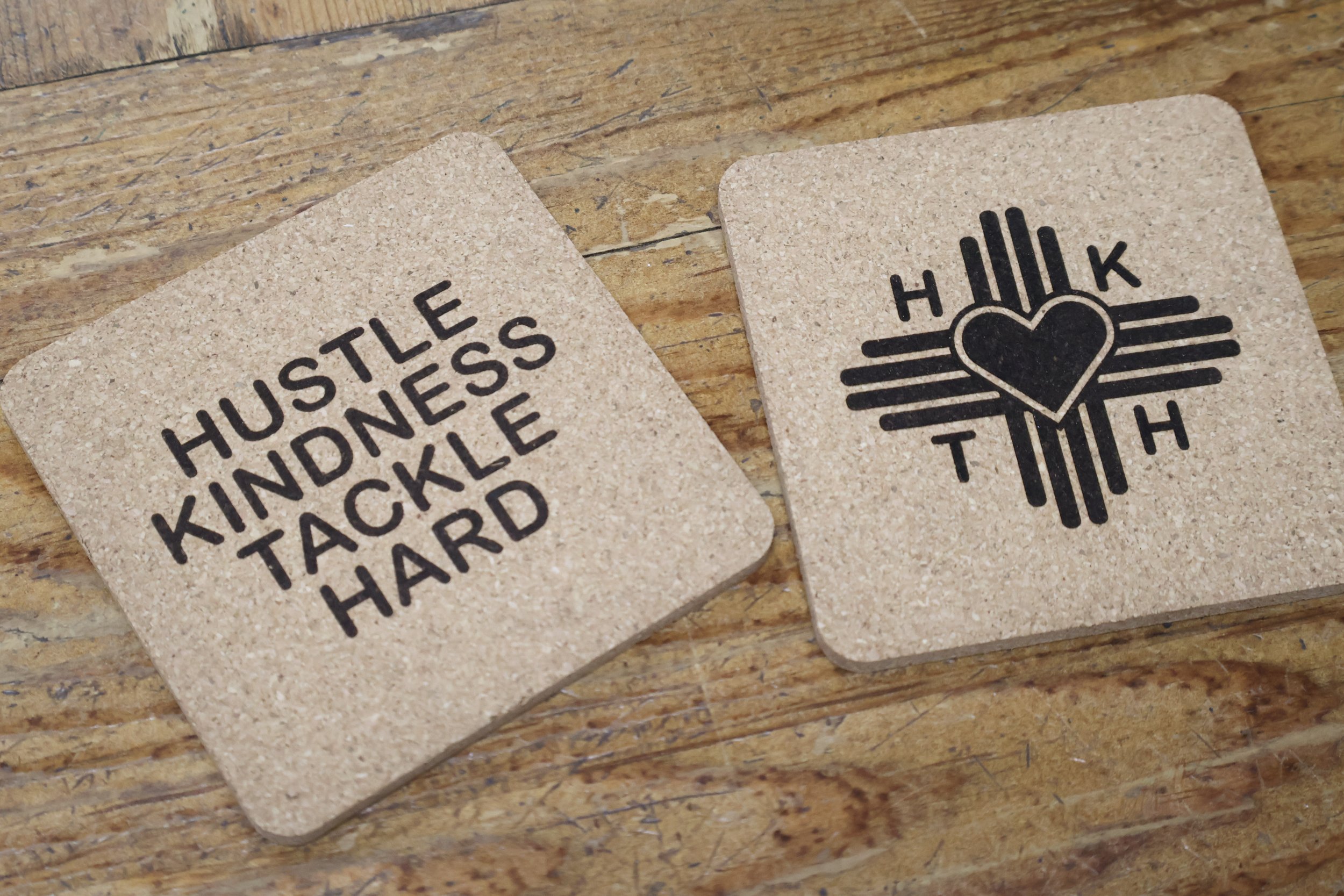 Two cork coasters on a wooden surface, one with the words "HUSTLE KINDNESS TACKLE HARD" and the other with a black graphic of a heart with lines radiating outward and the words "HOLD THEM".