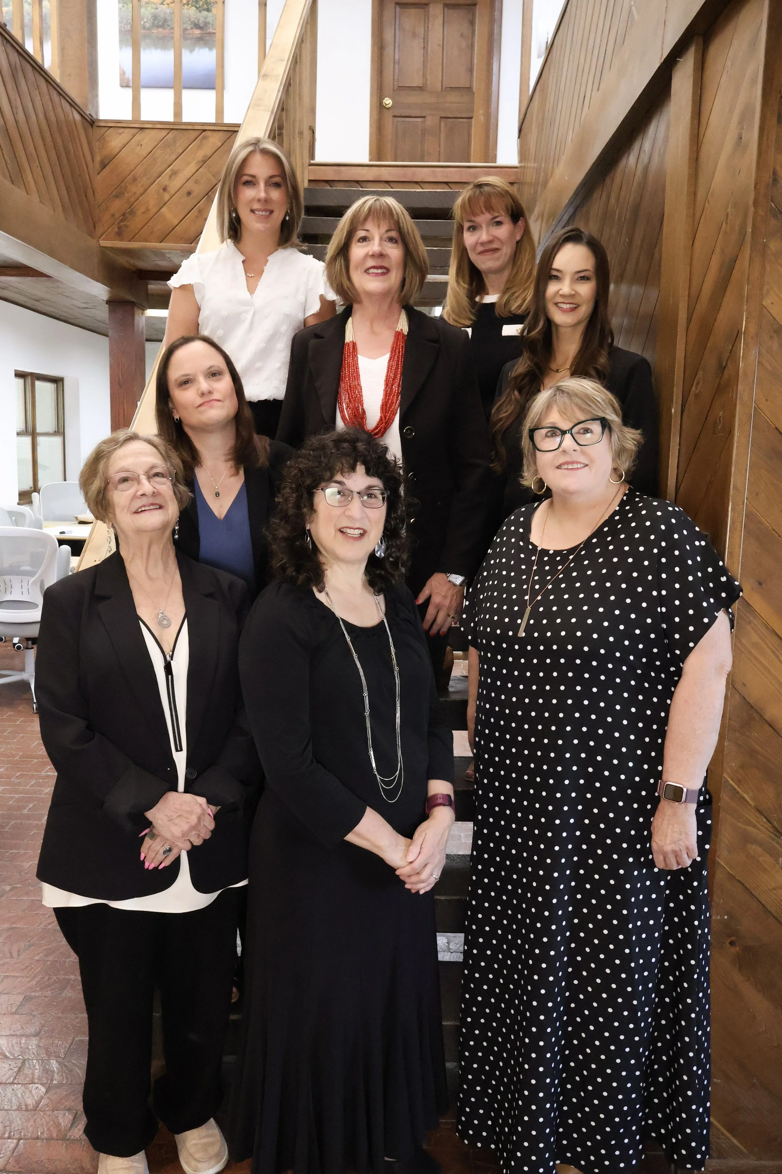 Group of nine women standing together inside a wooden staircase hallway, dressed in business and casual attire, smiling for the photo.