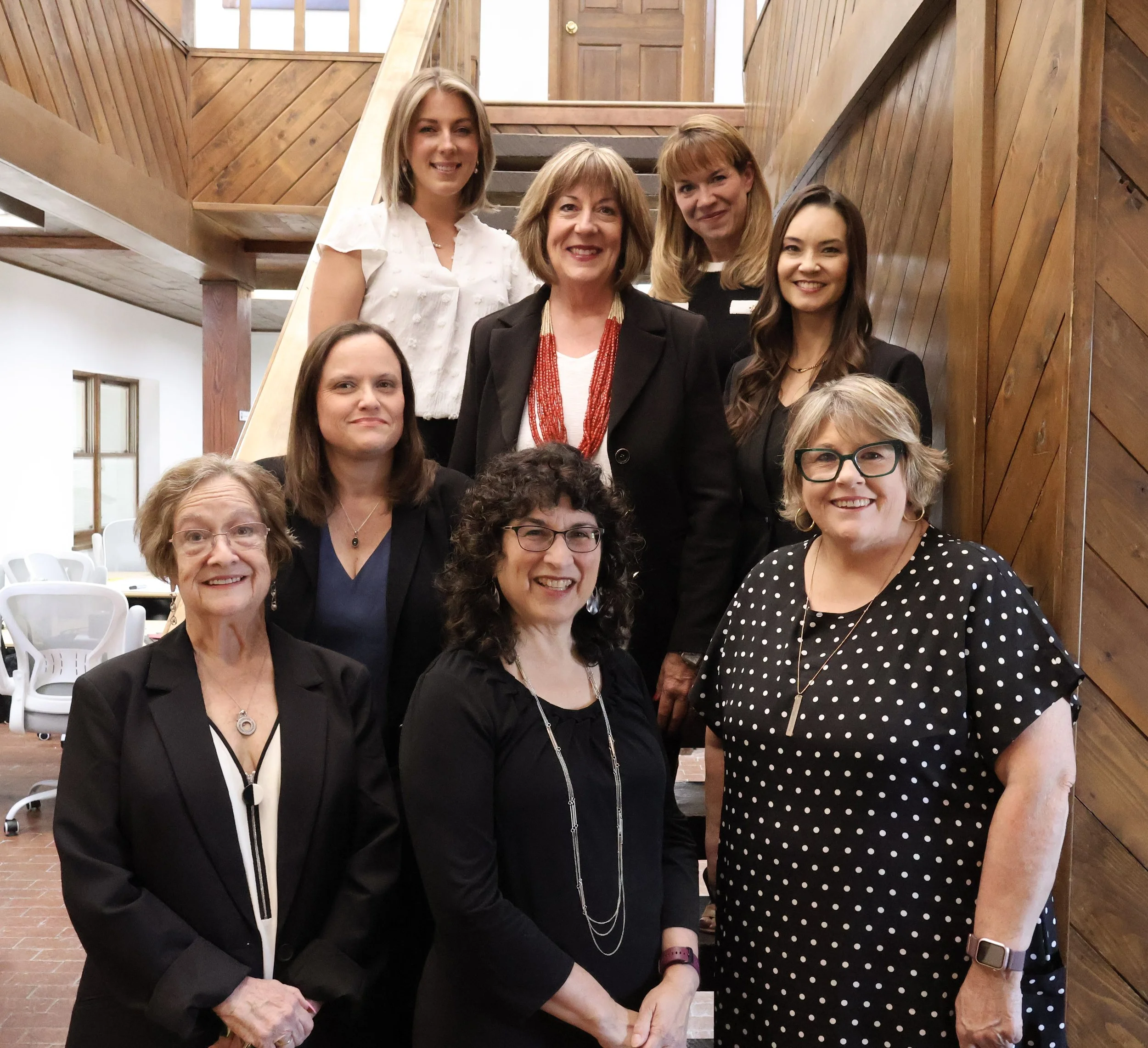 Group of women posing together indoors, with a wooden staircase and walls in the background.