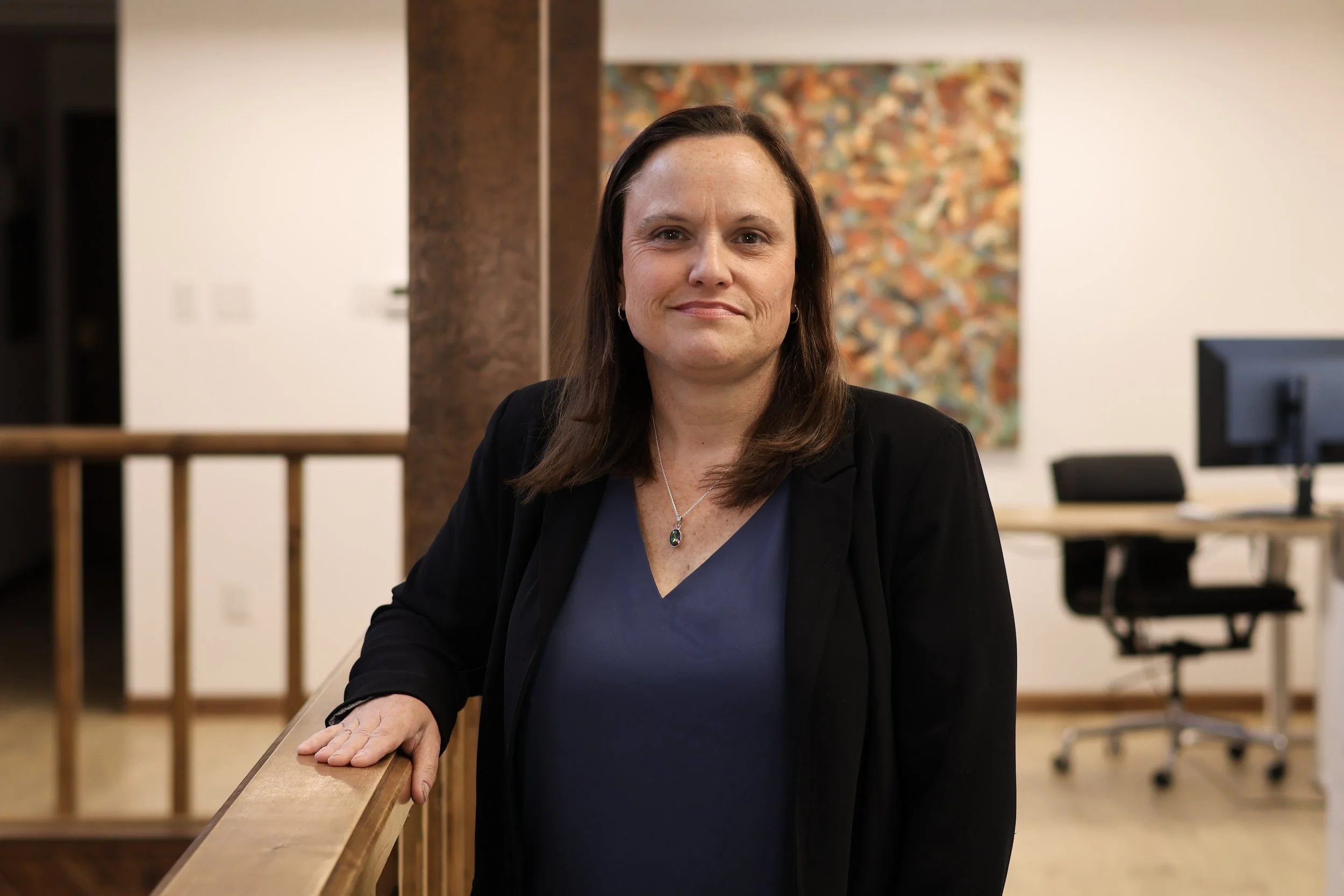 A woman with shoulder-length brown hair and wearing a black blazer and dark blue top, standing indoors with her hand resting on a wooden railing. There are chairs and a monitor in the background.