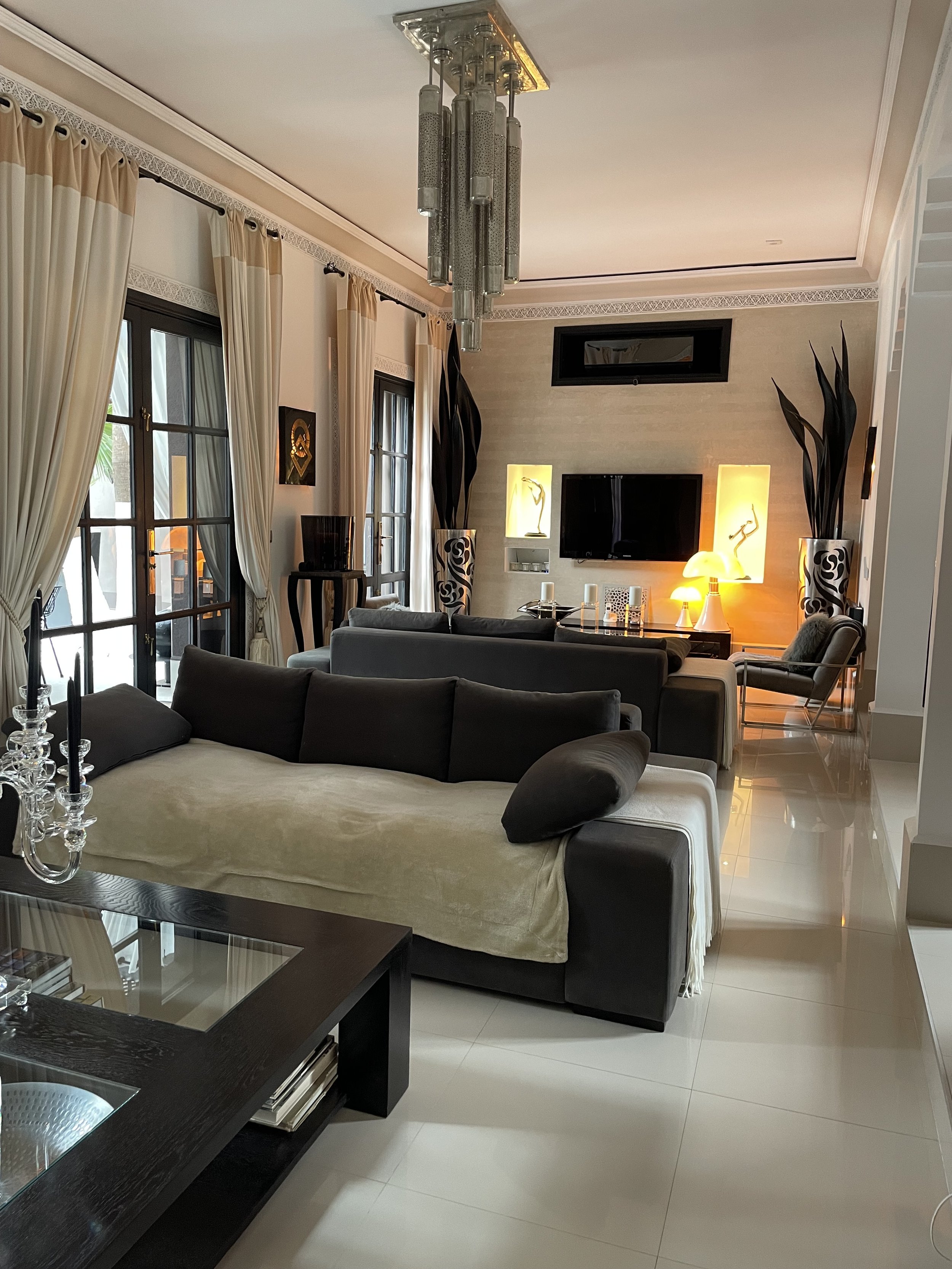 Salon moderne avec canapé en cuir noir, table basse en bois foncé avec surface en verre, grandes fenêtres avec rideaux beige, cheminée encastrée avec télévision, luminaires d'ambiance chaleureuse, plantes décoratives.