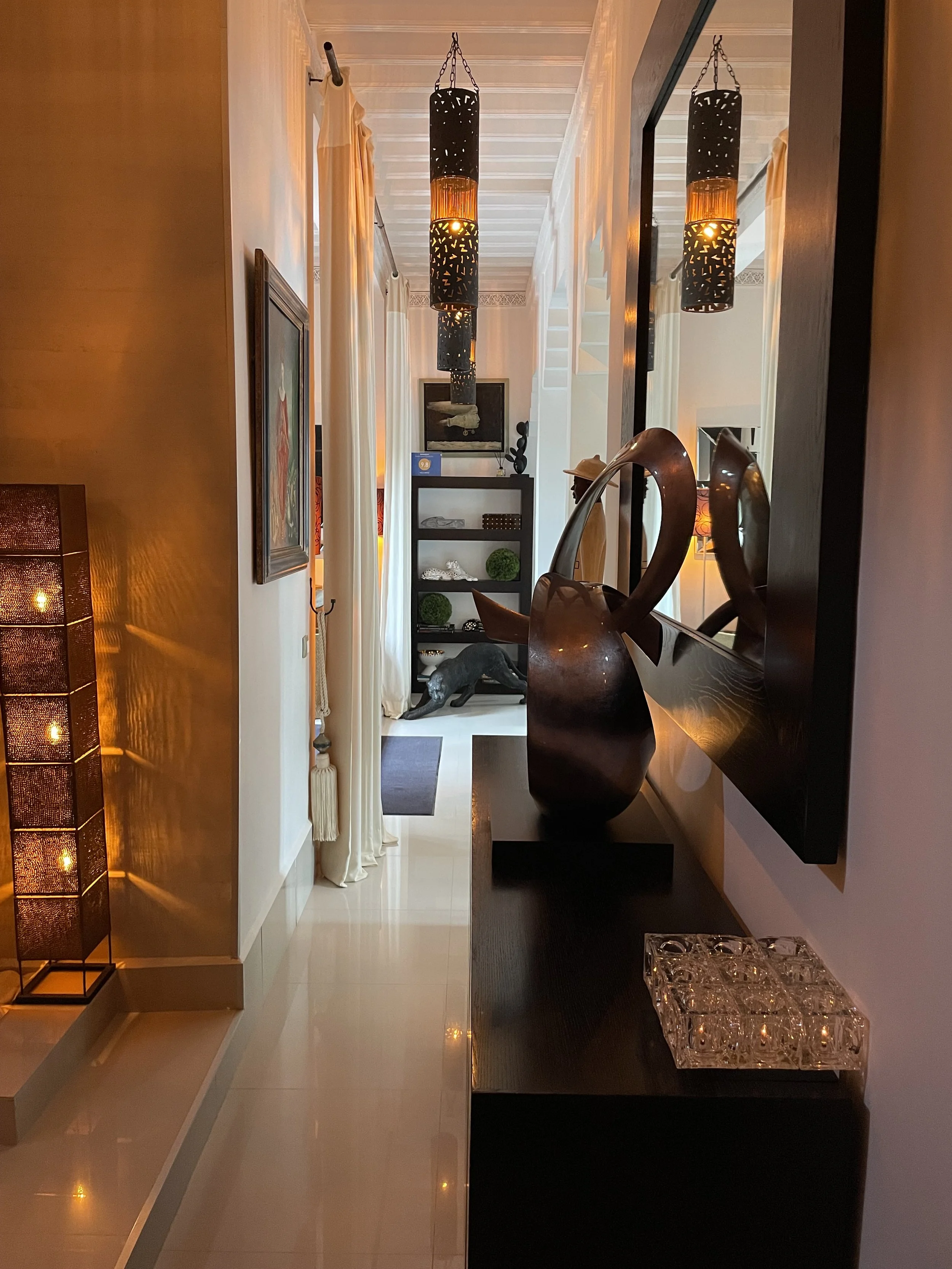 Vue d'un couloir intérieur avec des lampes suspendues design, un miroir, une étagère avec des objets décoratifs, et des rideaux blancs laissant passer la lumière naturelle.