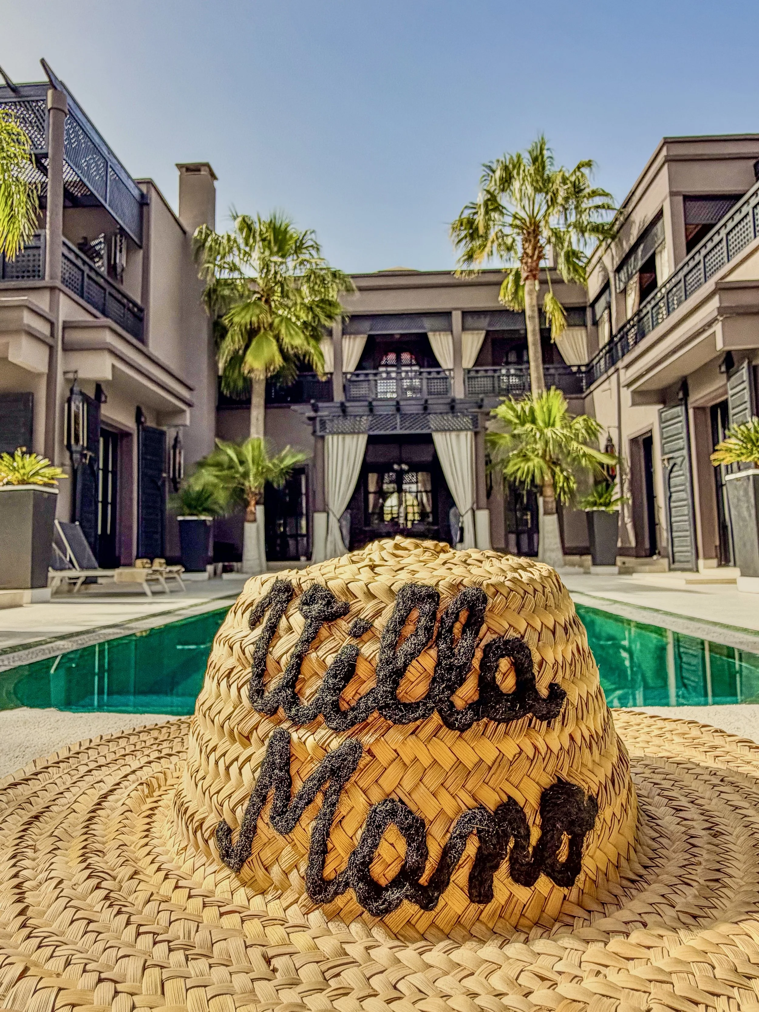 Chapeau de paille avec écriture noire en craie, posé à côté d'une piscine dans une cour d'hôtel avec palmiers et bâtiments modernistes