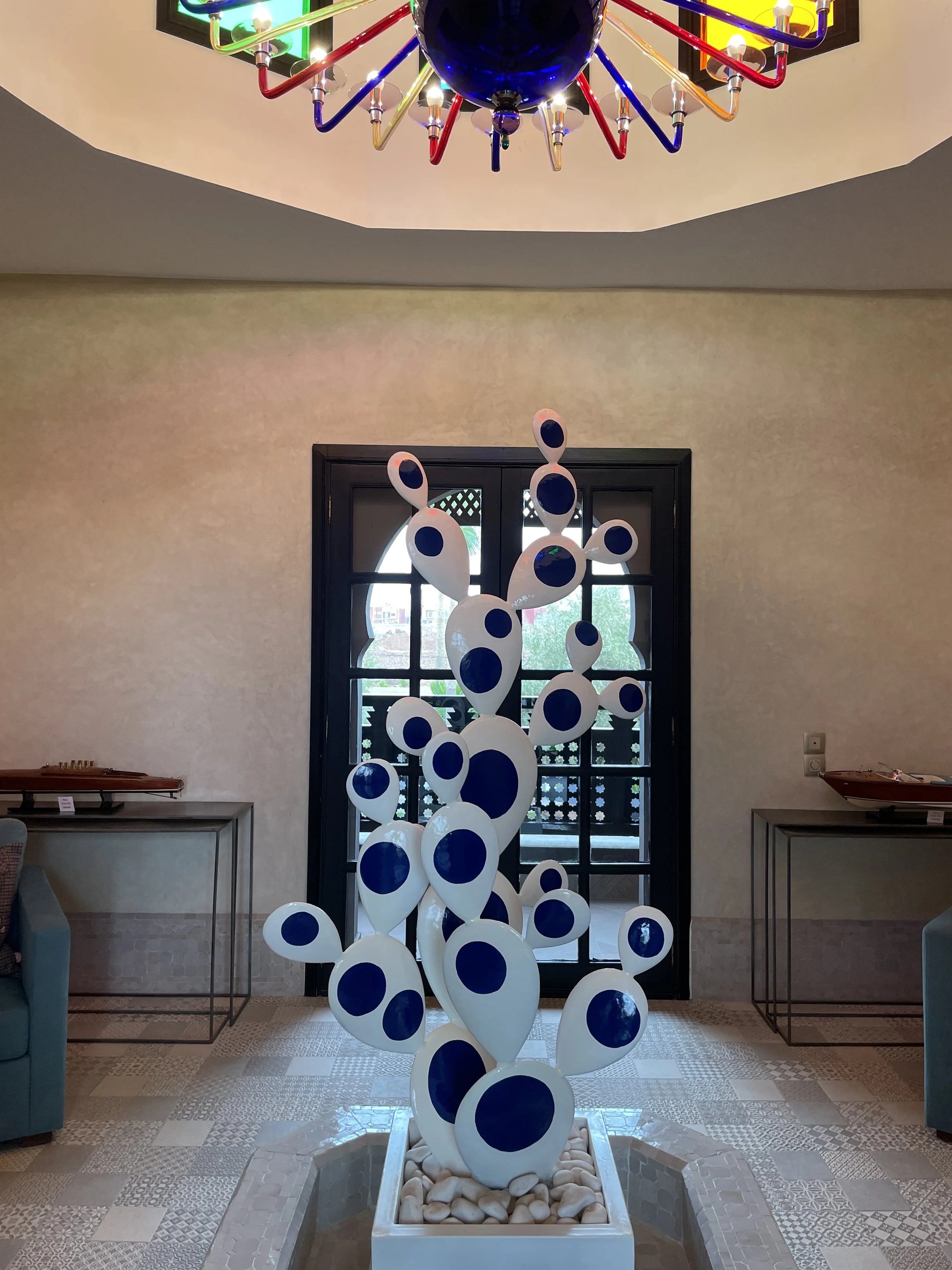 Une sculpture moderne de formant un cactus blanc avec des formes ovales et des cercles bleus, placée dans un vase rempli de pierres, dans une pièce avec une porte en verre et un plafond avec un lustre coloré.