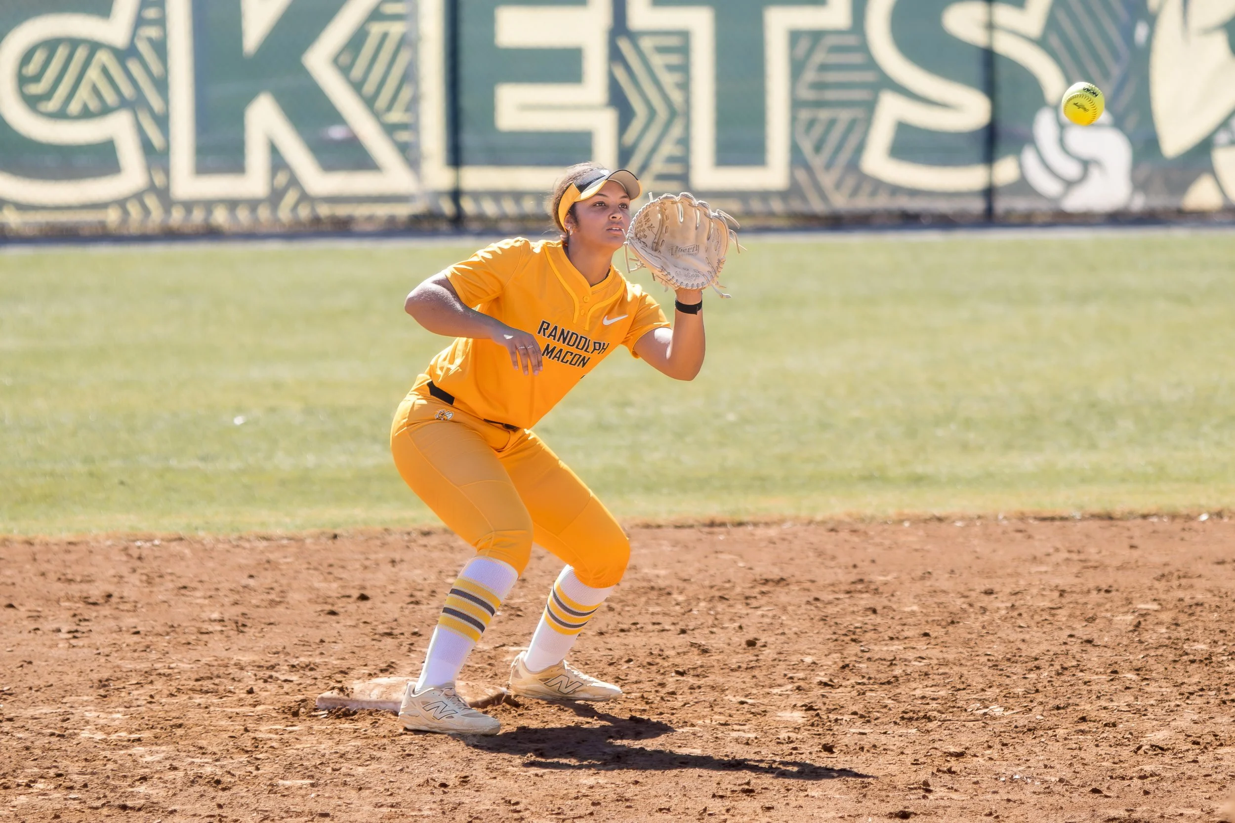 A female softball player in a yellow uniform is preparing to catch a yellow softball with black markings. She is on a dirt field with a grass outfield and is wearing a glove, white cleats, and striped socks, with a headband and a visor.