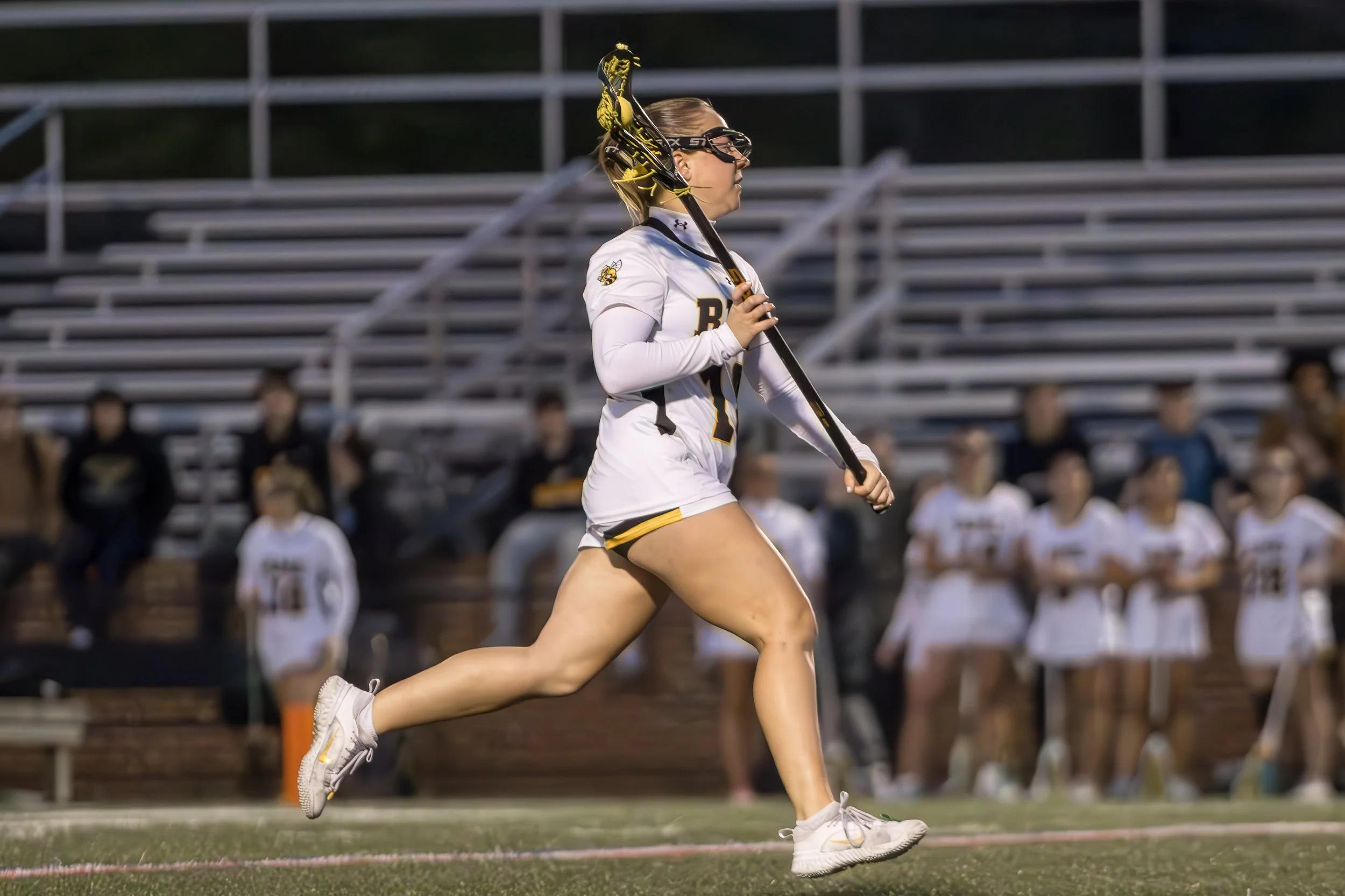 A female lacrosse player in a white and black uniform running on the field with a lacrosse stick, wearing goggles and a headband, with spectators in the bleachers in the background.