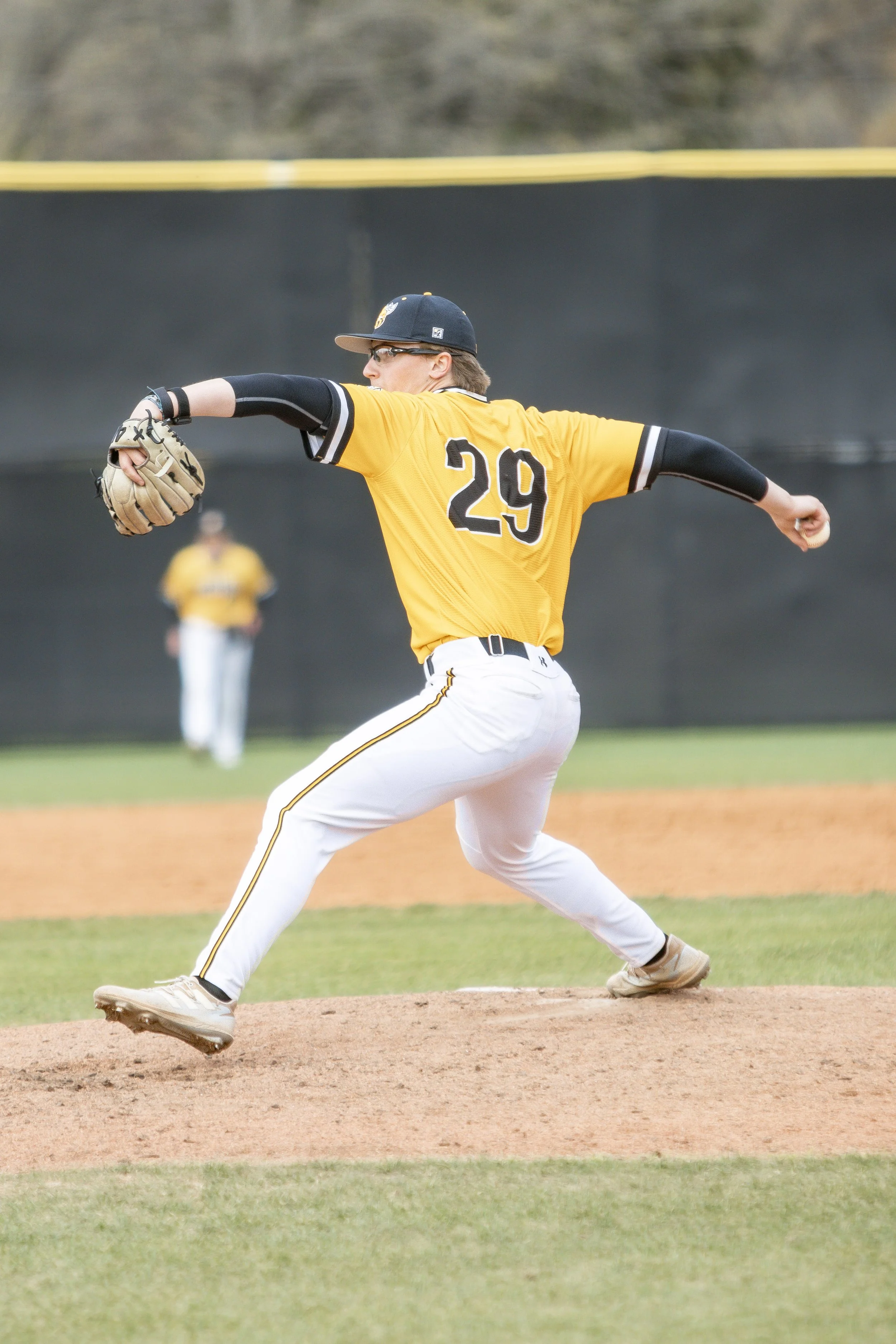 A baseball pitcher in a yellow uniform with the number 29 on the back, wearing a cap, glasses, and arm sleeves, pitching on the mound.