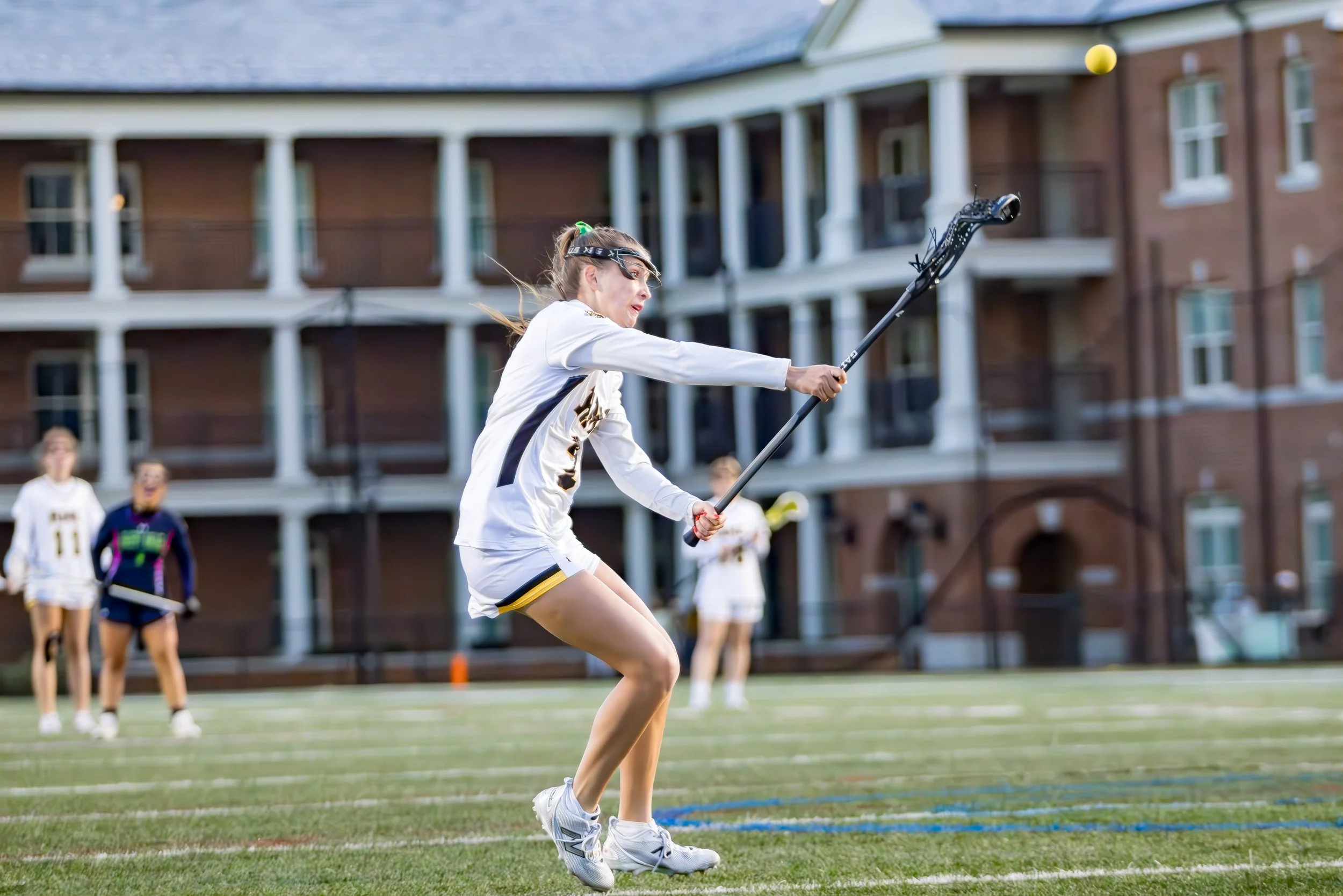 A female lacrosse player in white uniform is throwing a lacrosse ball with her stick on a green field, with other players and a brick building in the background.