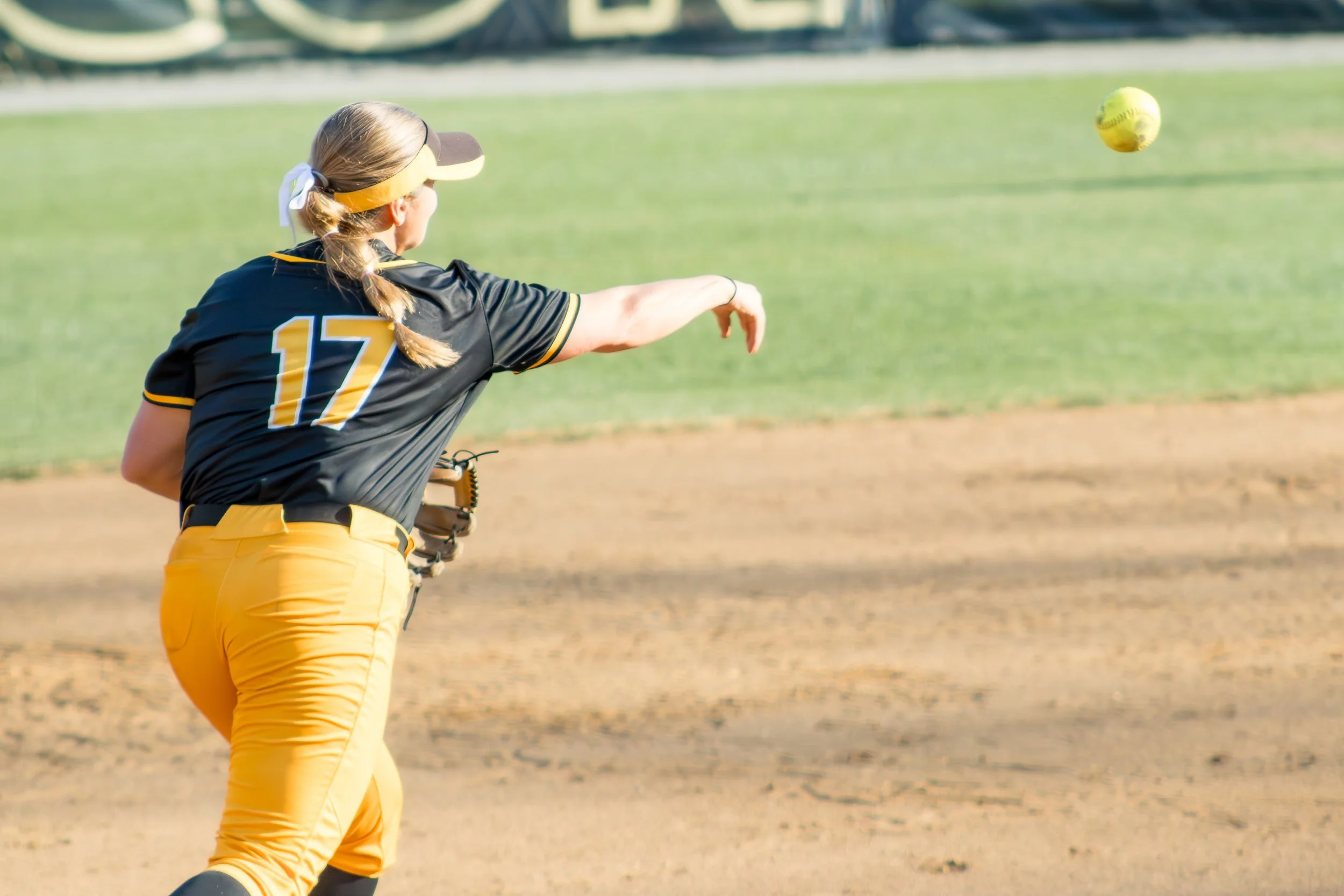 A female softball player wearing a black and yellow uniform with the number 17 on her back, in the act of throwing a yellow softball during a game on a dirt and grass field.