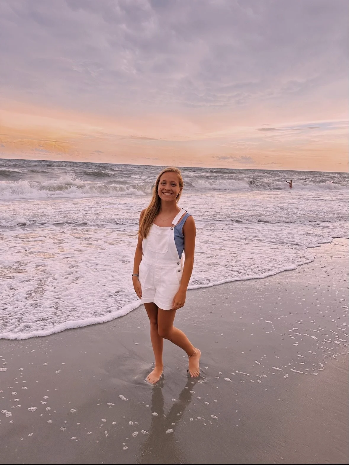 A young woman standing on the beach during sunset with the ocean in the background, smiling at the camera.